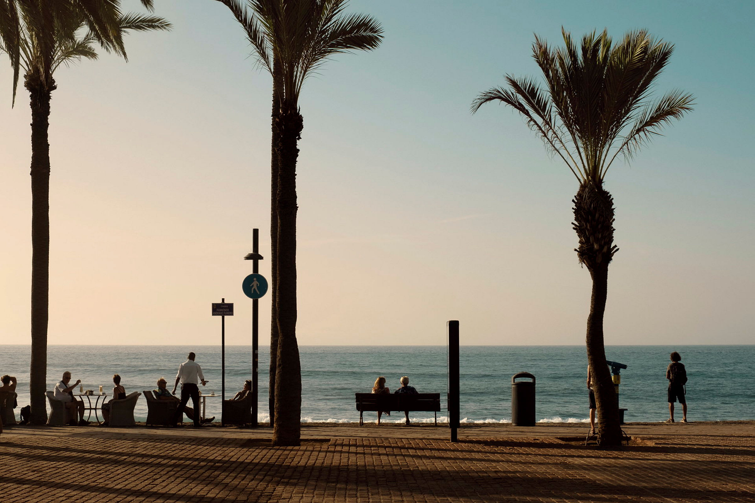 En strandpromenade med palmer, folk som sitter ved bord og ser mot havet. (AI-generert tekst)