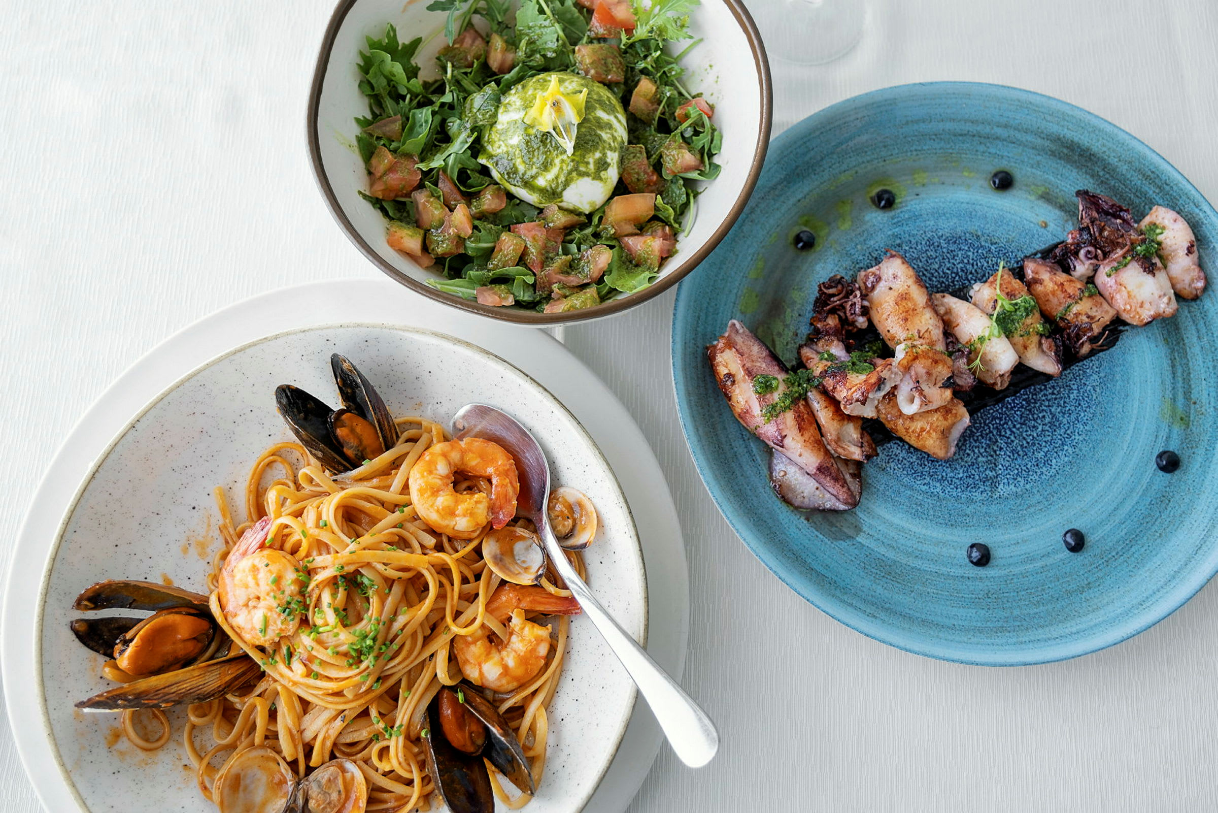 En tallerken med pasta og skalldyr, en salat med mozzarella og en tallerken med grillet blekksprut. (AI-generert tekst)