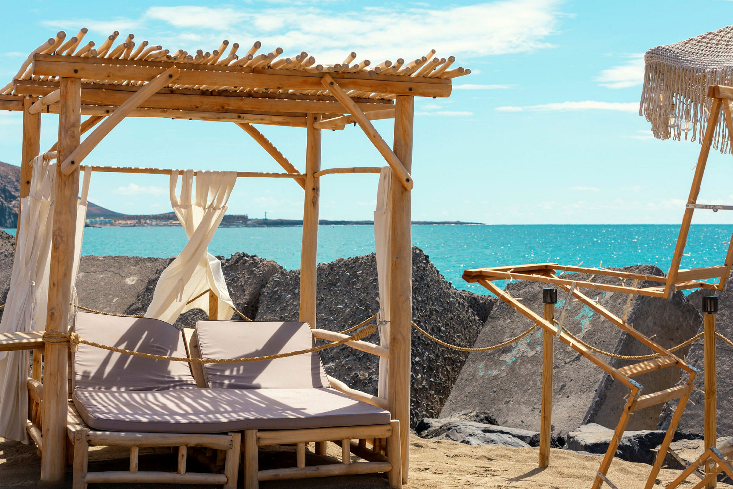 To solsenger under en trepergola på en sandstrand med utsikt over havet og store steinblokker. (AI-generert tekst)