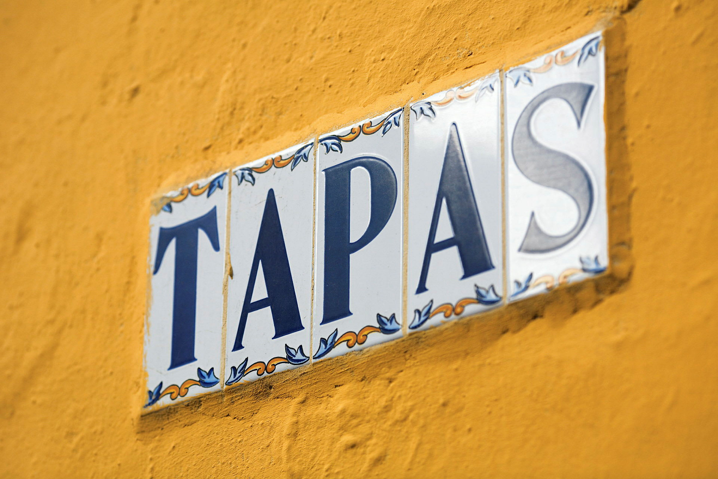 Et skilt med teksten 'TAPAS' på hvite keramiske fliser mot en gul vegg. (AI-generert tekst)