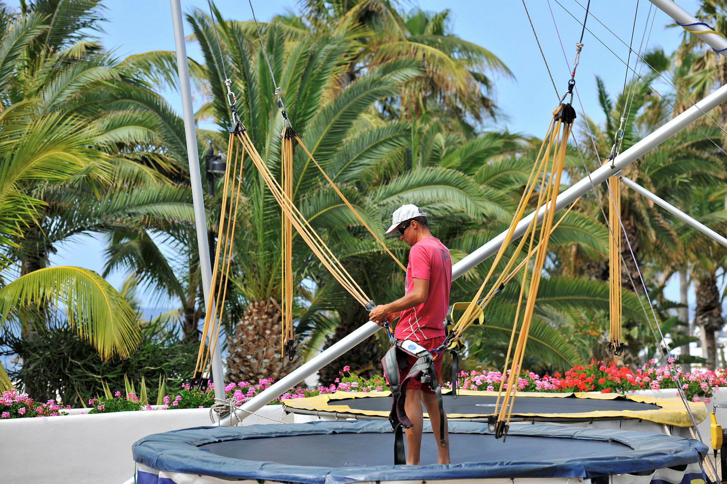 En person står ved en trampoline med sele og stropper. I bakgrunnen er det frodige palmetrær. (AI-generert tekst)
