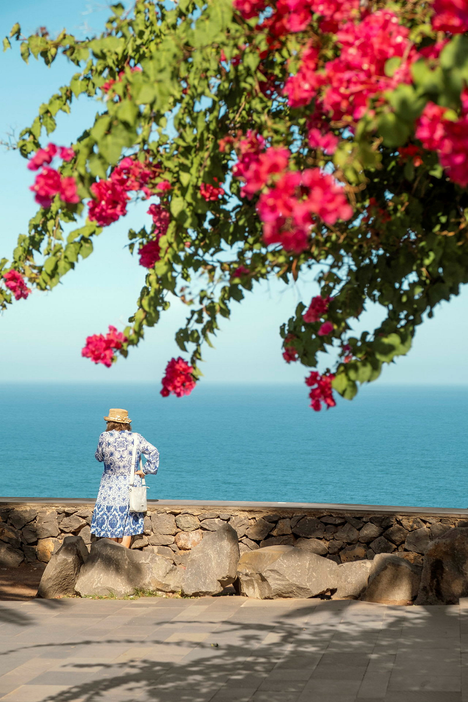 Person som står ved en steinmur med utsikt over havet under en blomstrende bougainvillea. (AI-generert tekst)