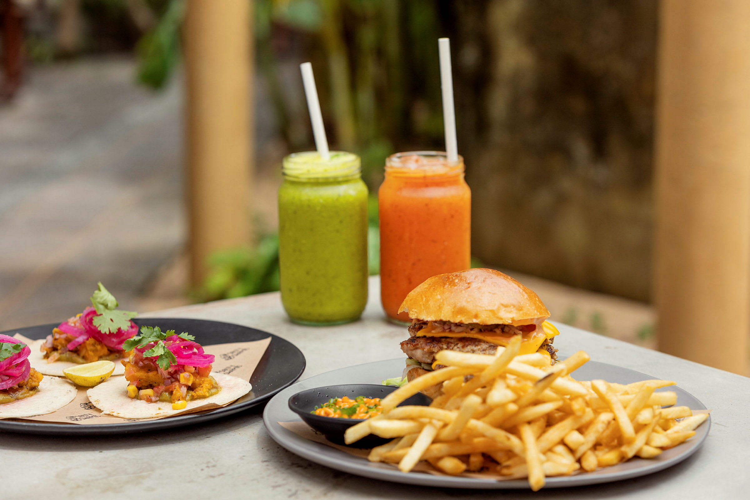 På bordet står en tallerken med tacos, en med hamburger og pommes frites samt to glass med fargerike drikker. (AI-generert tekst)