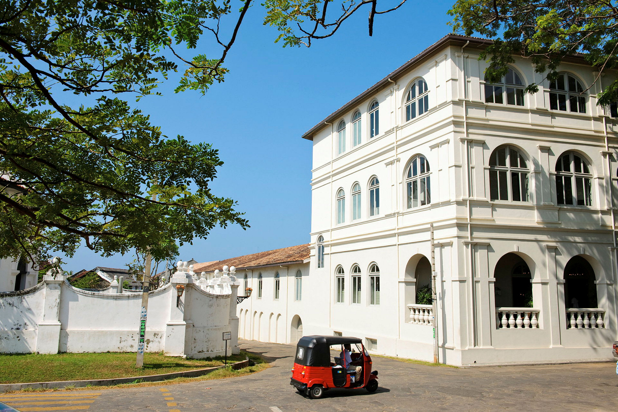 Et hvitt kolonialbygningskompleks med buede vinduer og detaljer. Foran bygningen står en rød tuk-tuk parkert, omgitt av grøntområder under en klar blå himmel. (AI-generert tekst)