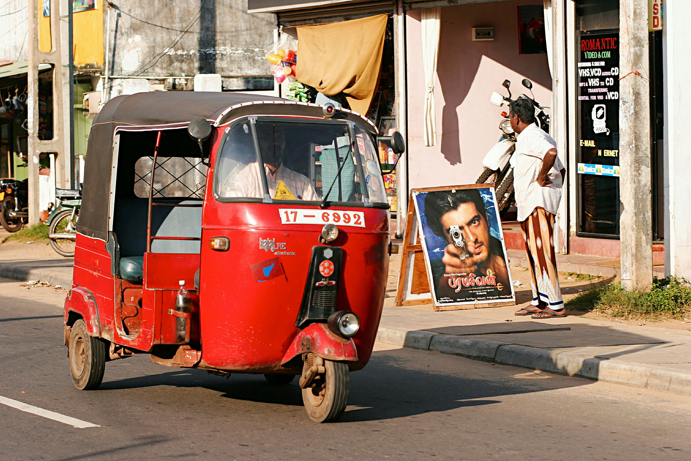 En rød tuk-tuk kjører forbi en butikk på en solrik gate. (AI-generert tekst)
