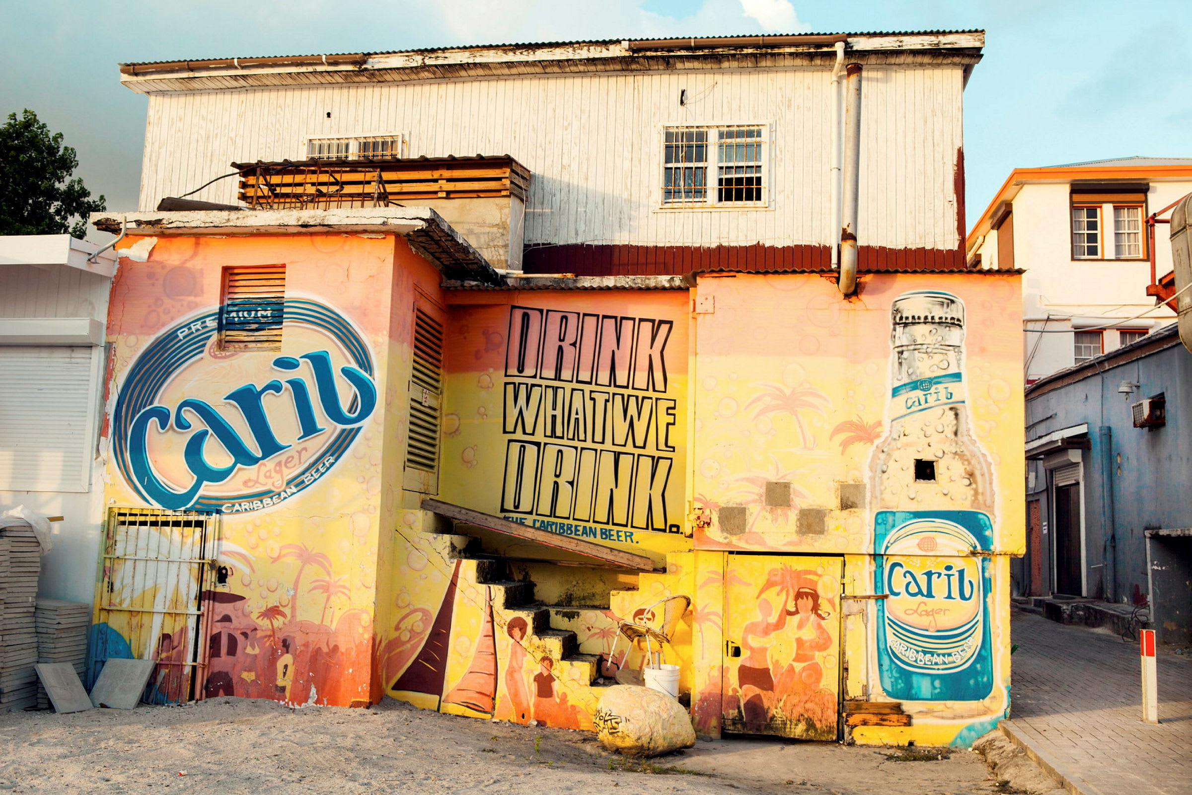 En fargerik husvegg malt med reklame for Carib-øl, inkludert et stort bilde av en Carib-flaske og teksten 'Drink what we drink'. (AI-generert tekst)