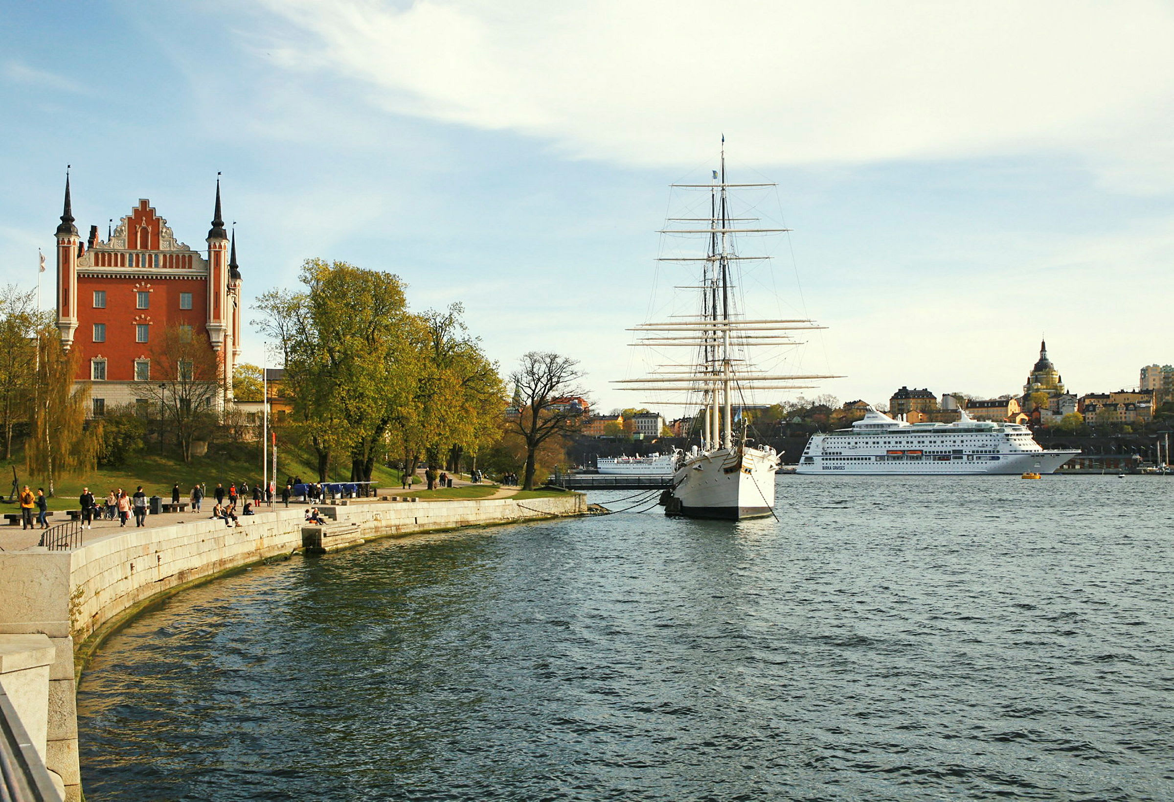 Et rødt hus med tårn ved en brygge, et seilskip og et cruiseskip sees ved vannet. (AI-generert tekst)