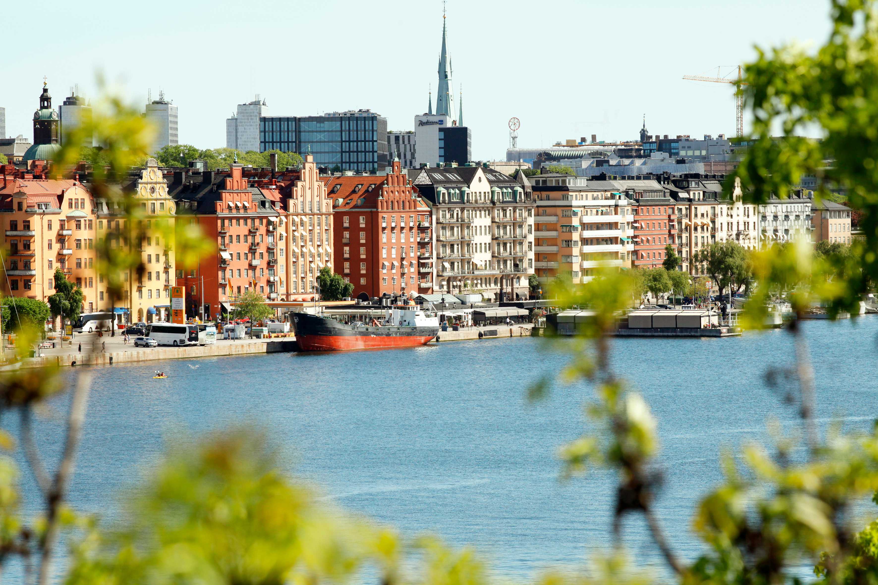 Utsikt over en strandlinje med bygninger og båter ved vannet, delvis innrammet av grøntområde. (AI-generert tekst)