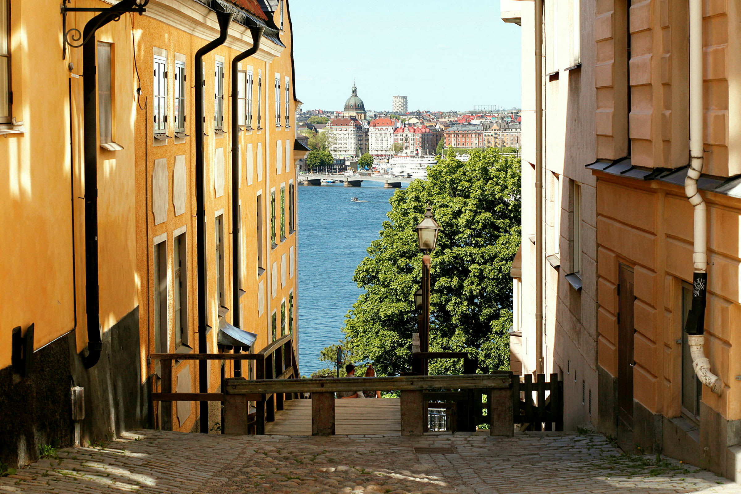Et gatebilde med oransjefargede bygninger på hver side, som skråner ned mot vann og en by i bakgrunnen. (AI-generert tekst)