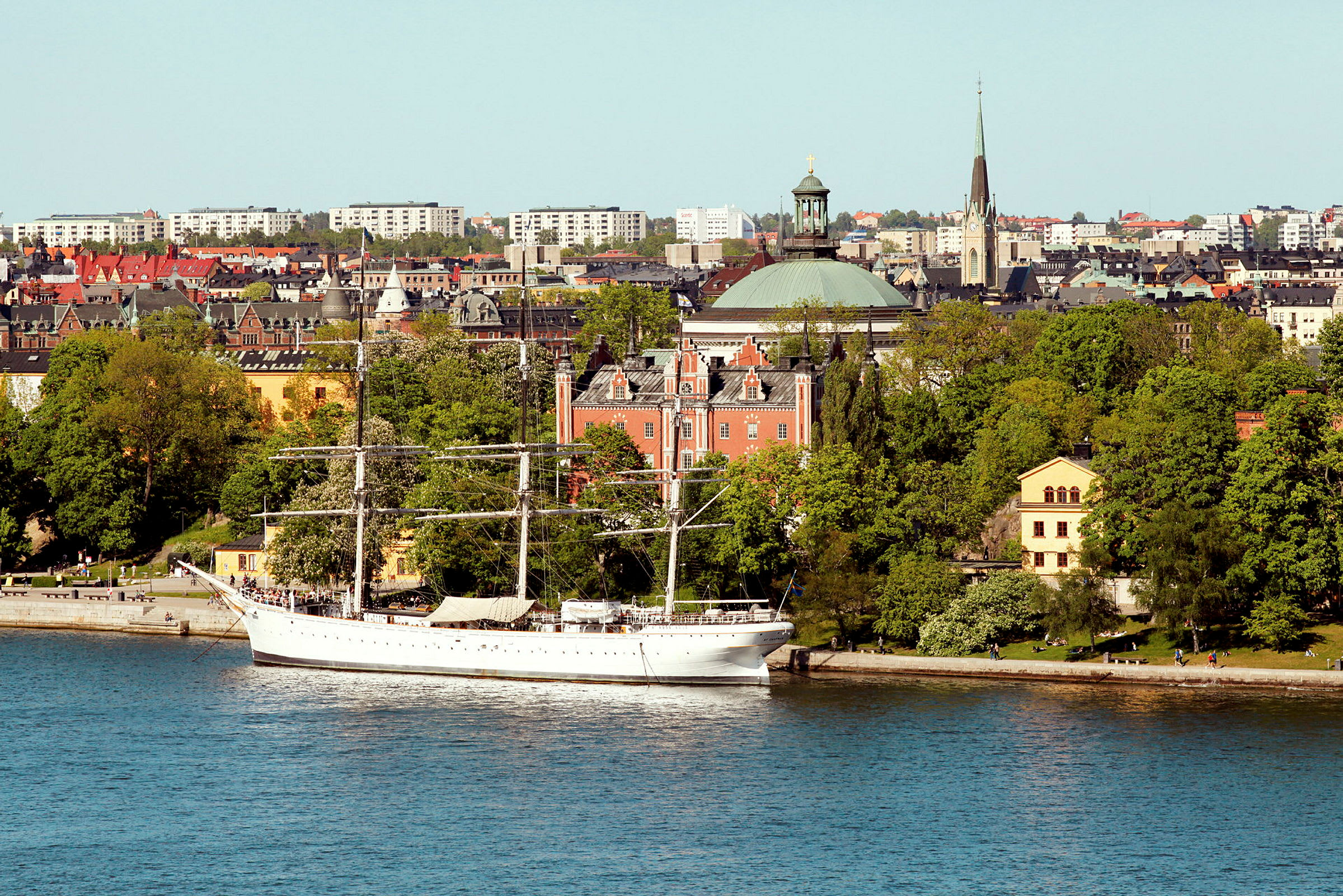 Et hvitt seilskip ligger for anker ved en kai i en by ved vannet. Bakgrunnen består av trær, flere eldre bygninger og bygningsstrukturer. (AI-generert tekst)