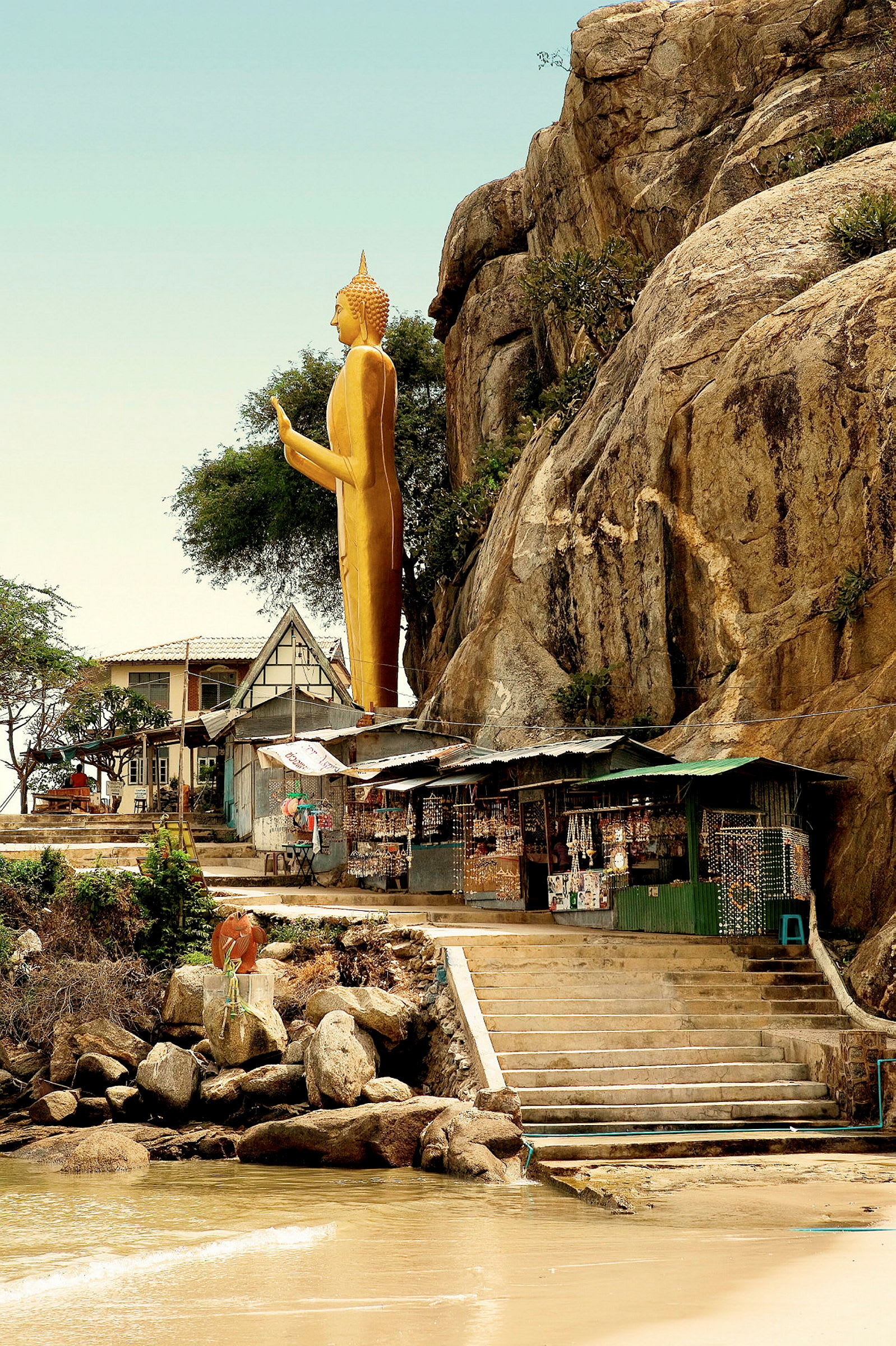 En gylden buddhastatue står ved en klippe over en liten marked og trapper som fører ned til stranden. (AI-generert tekst)