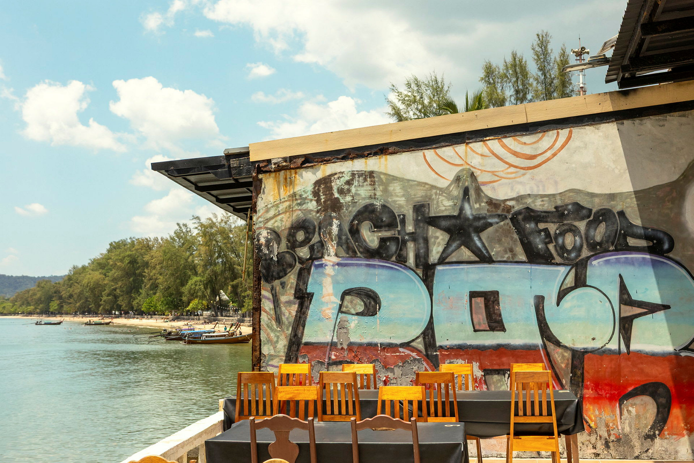 En utsikt over en strand med vann, trær og båter. Foran i bildet sees en vegg med graffiti samt stoler og bord. (AI-generert tekst)
