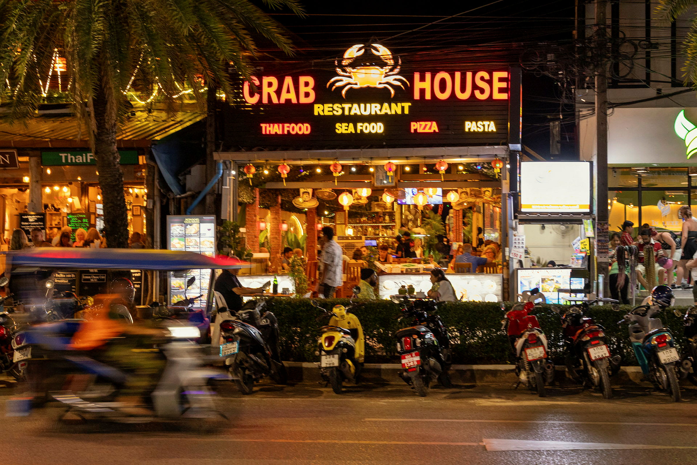 Bilde av en livlig gate med restauranten Crab House i fokus. Flere motorsykler står parkert foran restauranten, og en motorsykkel med en passasjer kjører forbi. (AI-generert tekst)