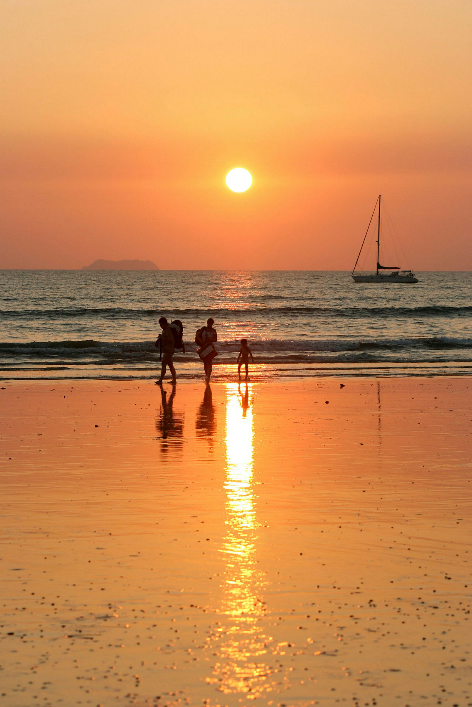 Fire personer går på stranden ved solnedgang, med en seilbåt synlig på havet. (AI-generert tekst)