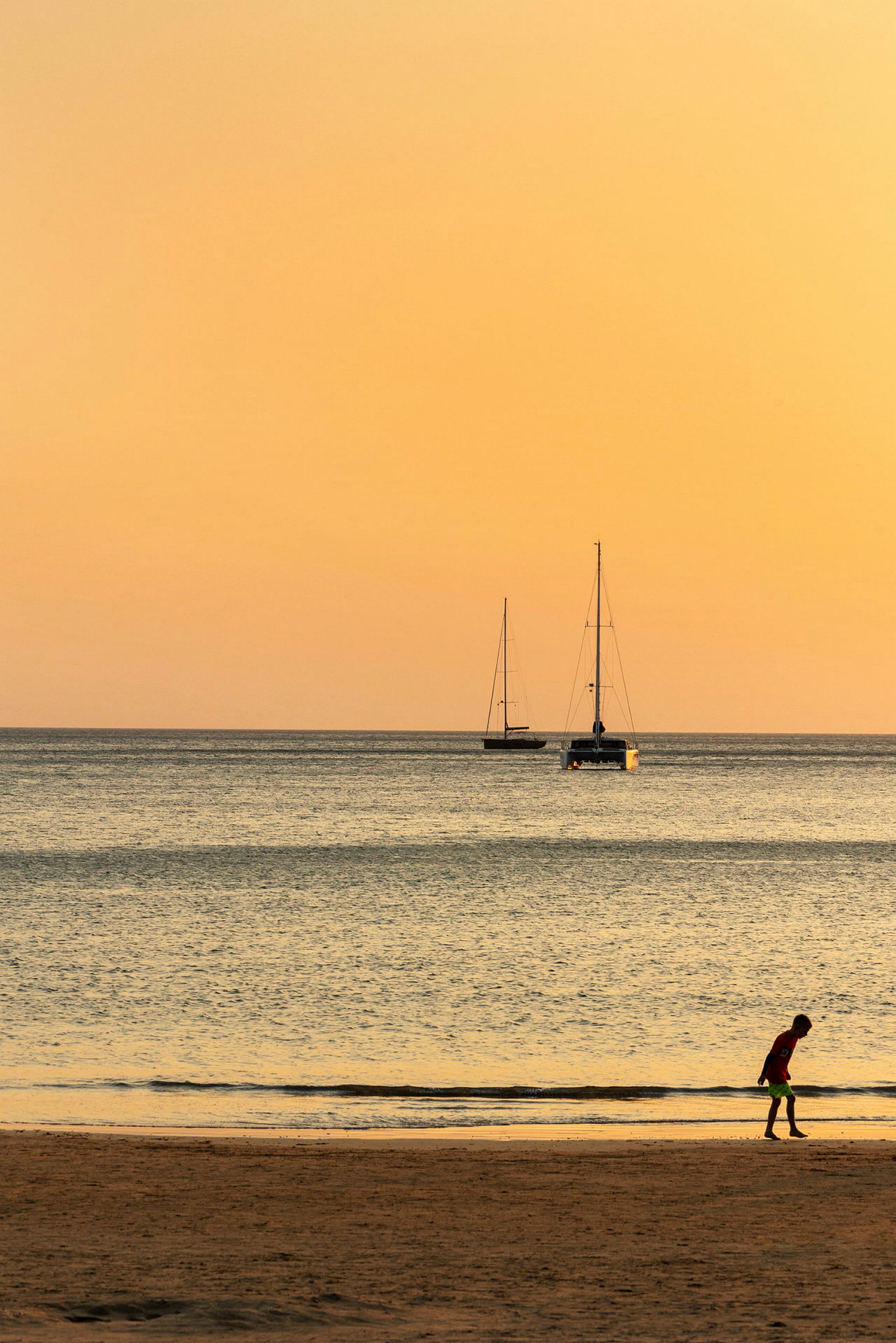 En strand ved havet under en oransje solnedgang med to seilbåter i bakgrunnen og en person som går i sanden. (AI-generert tekst)