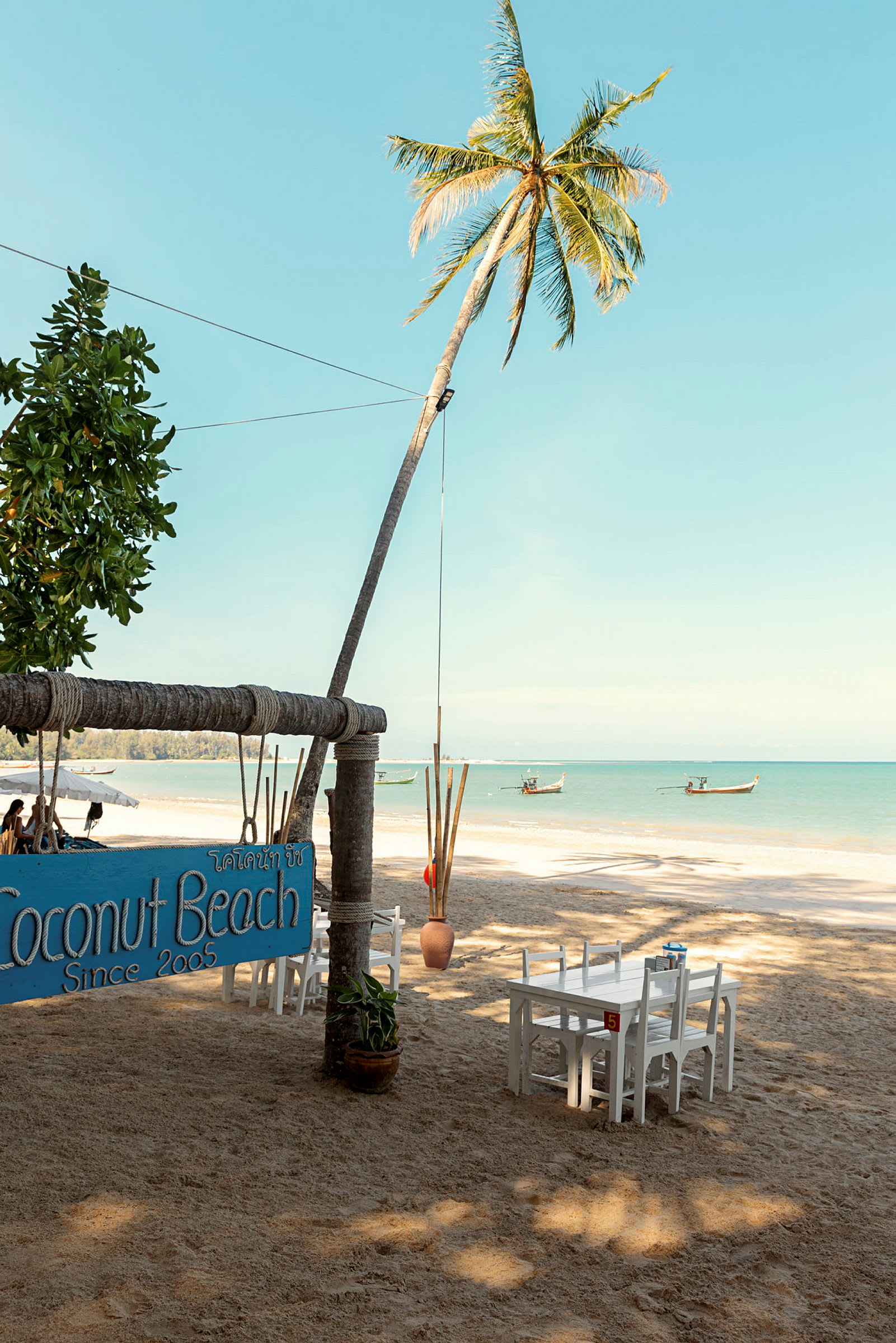 En strand med palmer, bord og stoler, samt en skilt som markerer Coconut Beach. (AI-generert tekst)