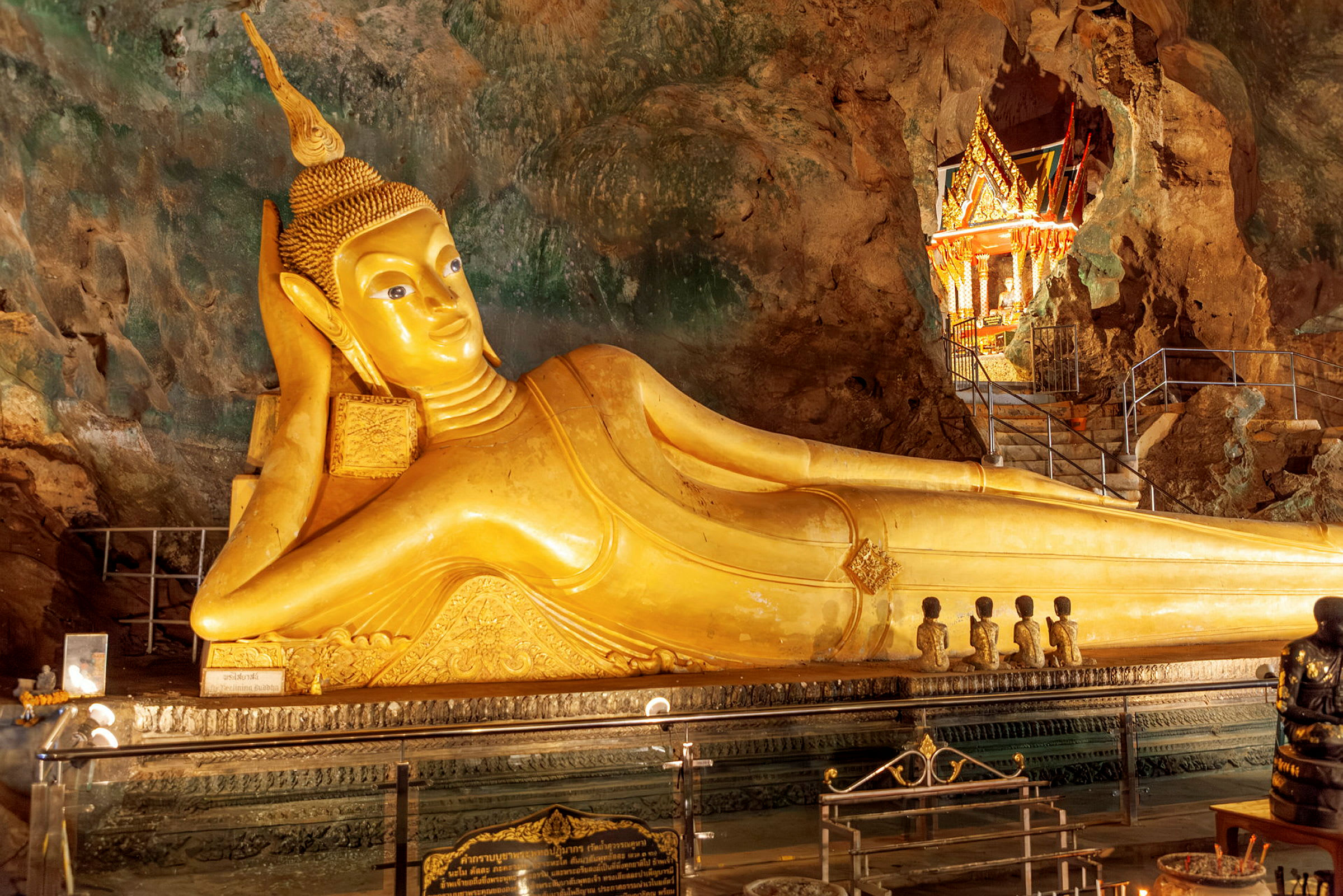 En gyllenliggende Buddha-statue inne i en grotte, omgitt av et vakkert dekorert område med detaljerte utsmykninger. (AI-generert tekst)