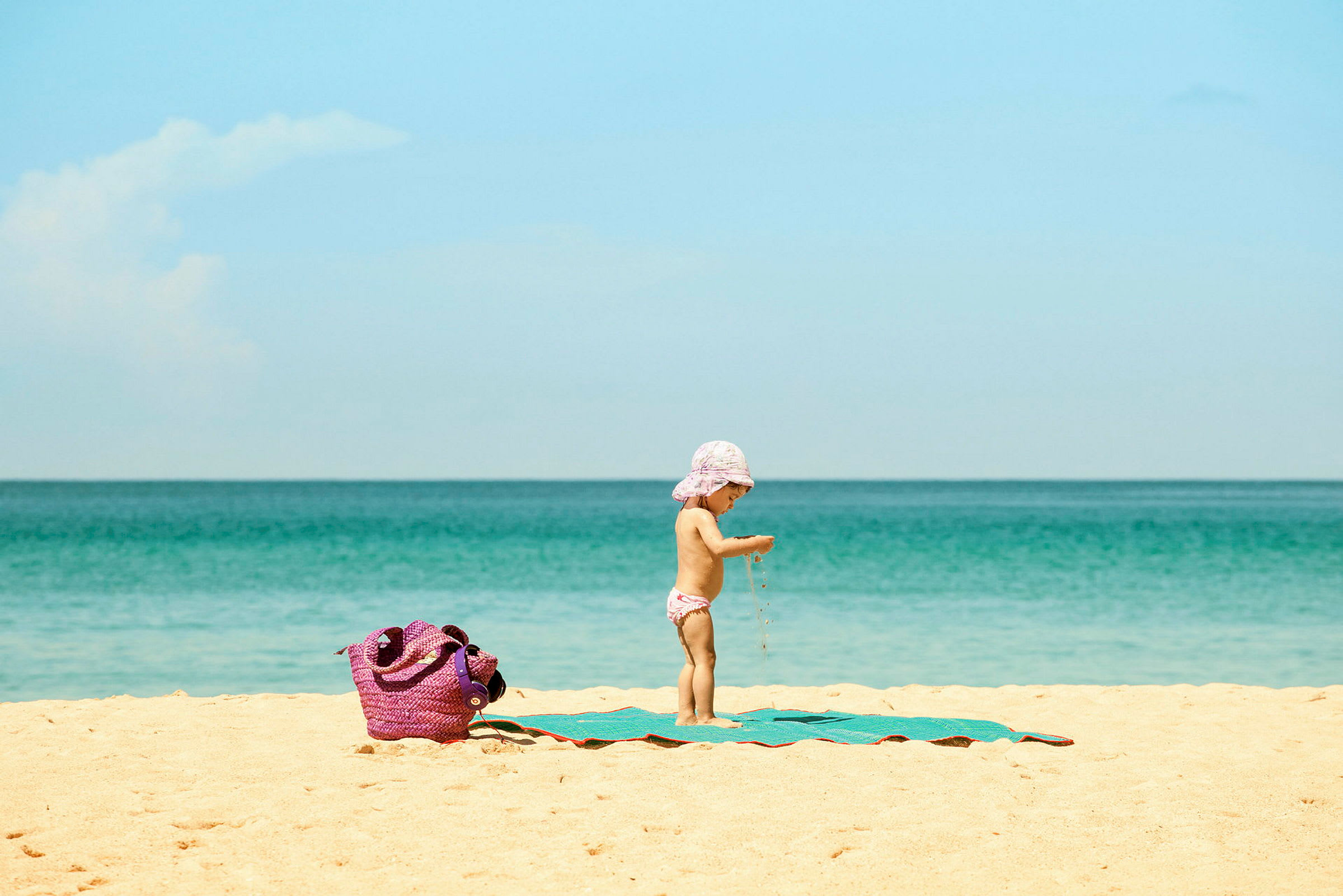 Et barn på en strand med et hav i bakgrunnen og en lilla veske på sanden. (AI-generert tekst)