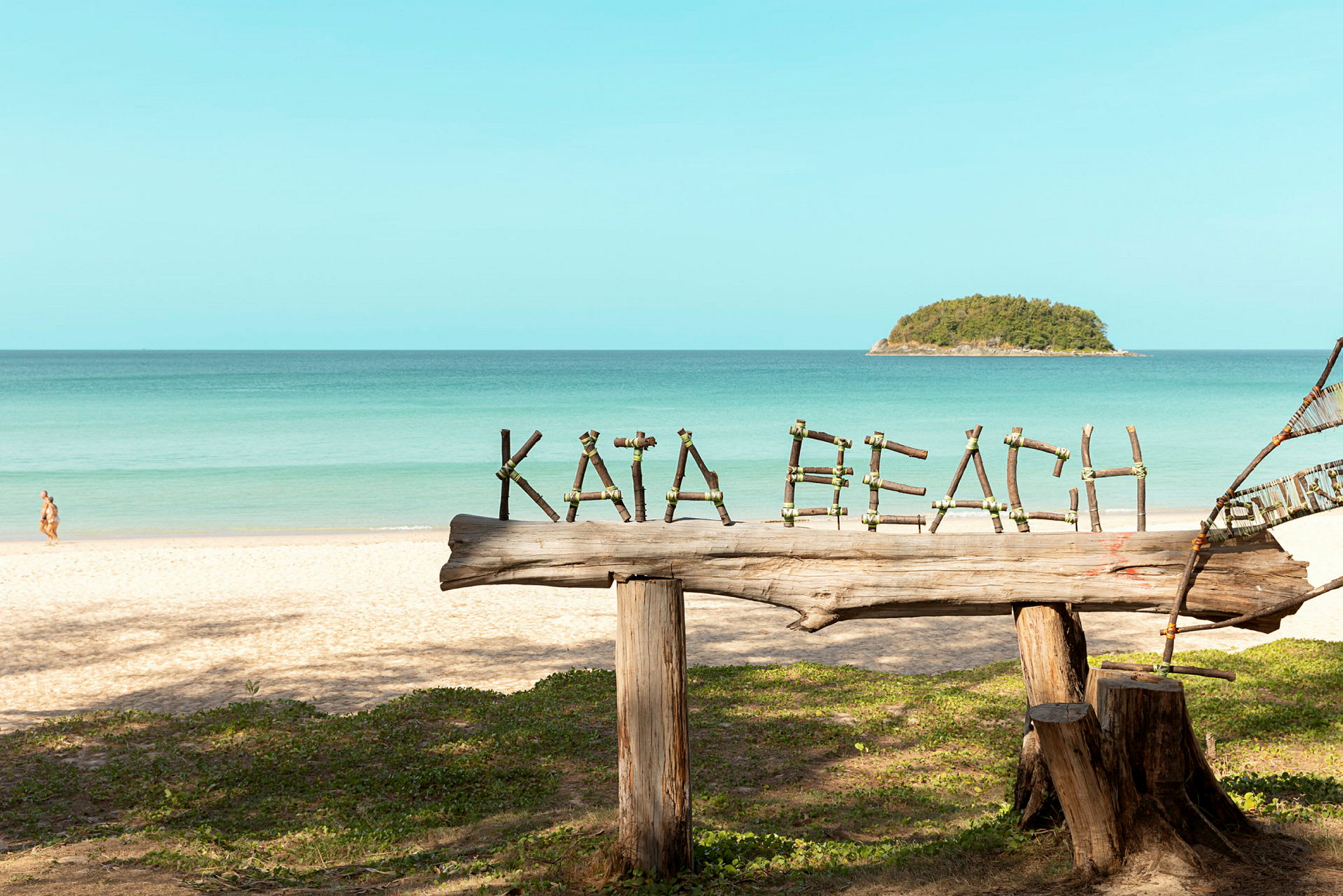 Treskilt med teksten 'Kata Beach' på en sandstrand med havet i bakgrunnen. (AI-generert tekst)