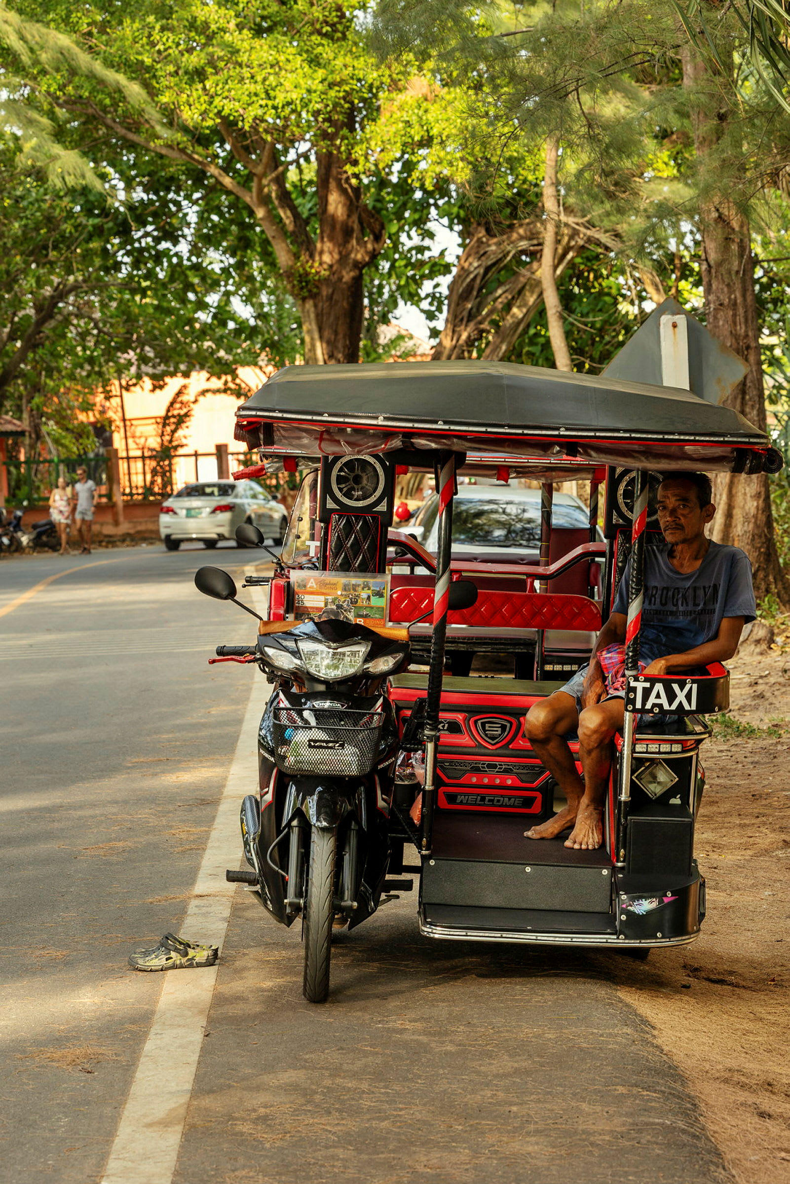 En tuk-tuk står parkert ved siden av en vei. En mann sitter i kjøretøyet og omgivelsene består av trær og noen parkerte biler. (AI-generert tekst)