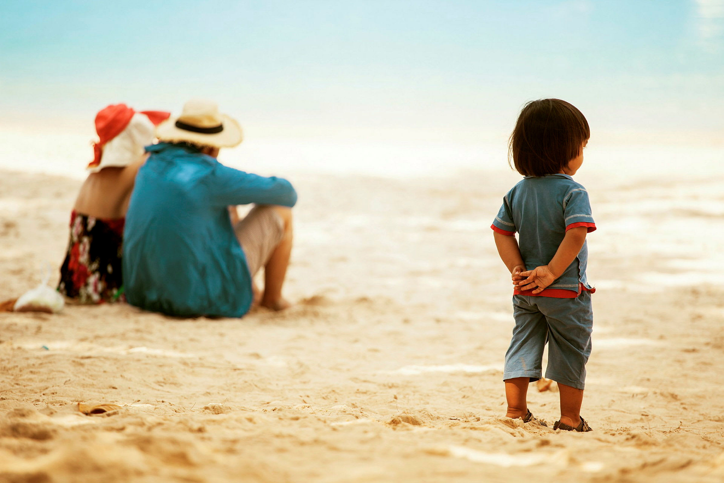 Et barn står på stranden med hendene bak ryggen, mens to voksne sitter lengre bort. (AI-generert tekst)