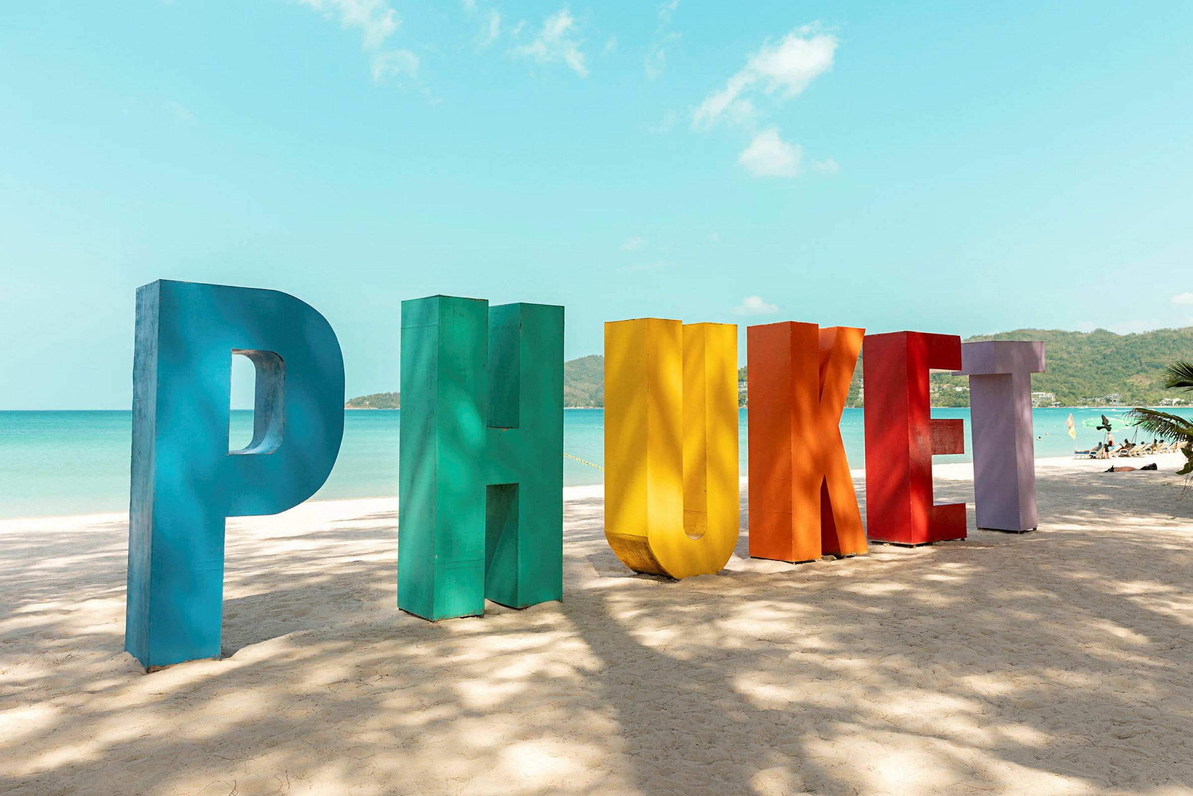 Fargerike bokstaver danner ordet 'Phuket' på en sandstrand, med hav og himmel i bakgrunnen. (AI-generert tekst)