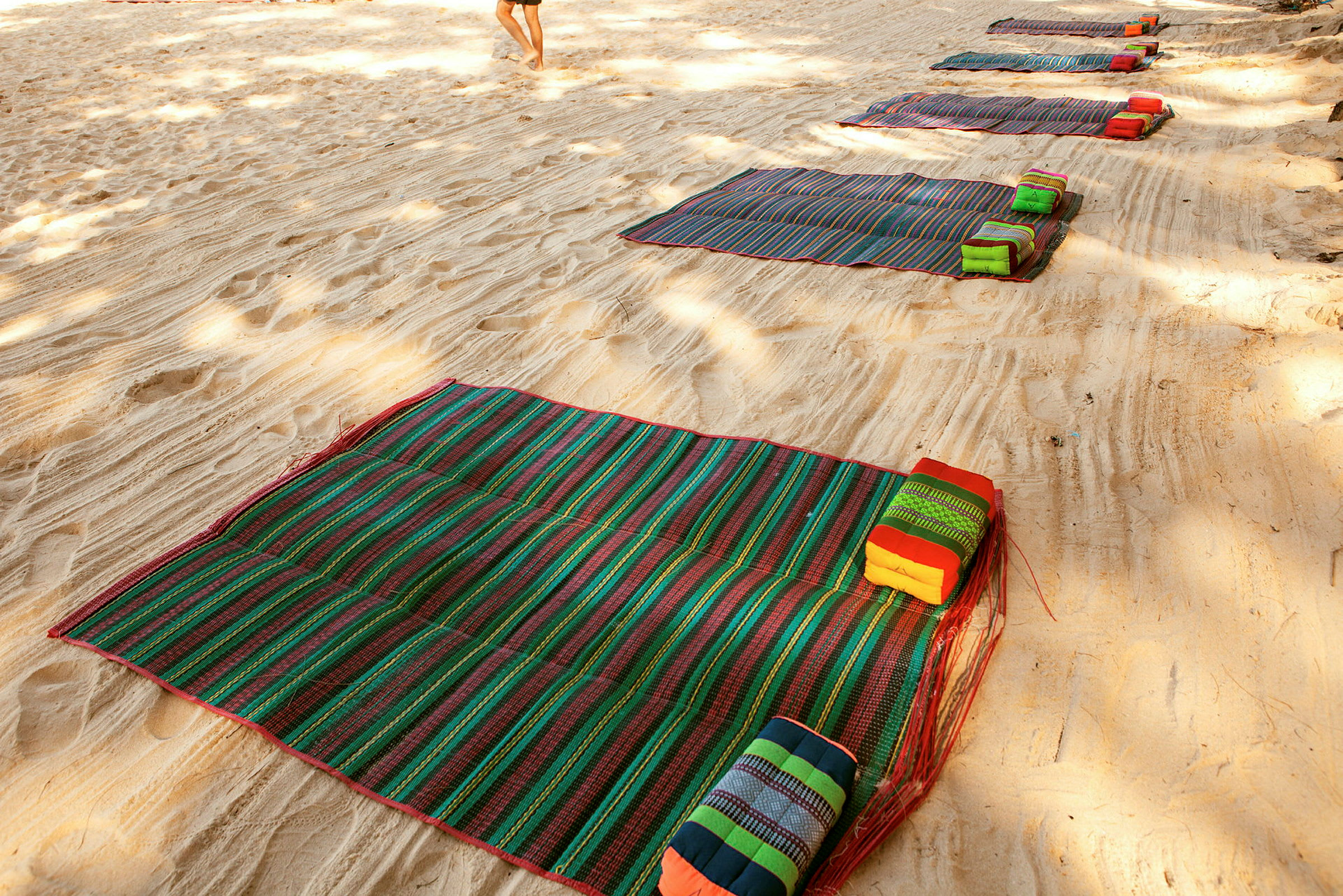Flere fargerike strandmatter med puter plassert på sandstrand. (AI-generert tekst)