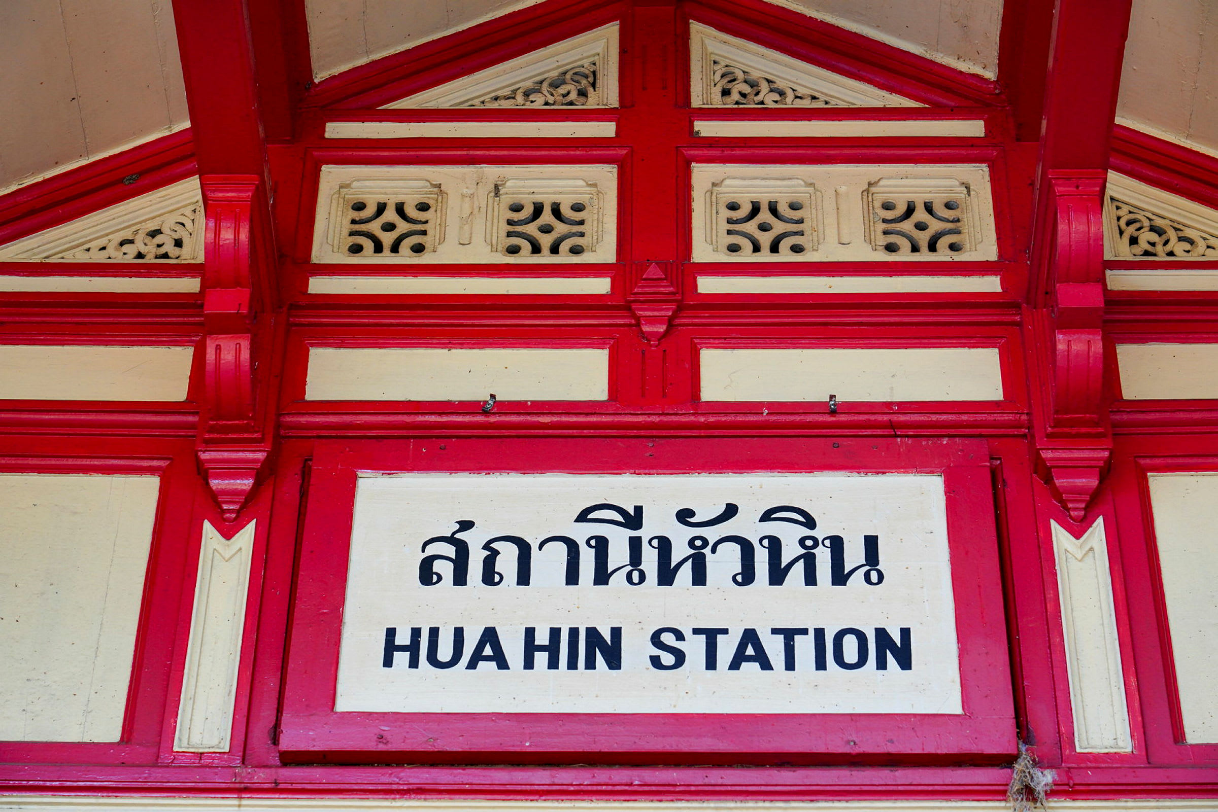 Skilt med teksten 'Hua Hin station' med thailandsk skrift og dekorativt rød og hvitt rammeverk. (AI-generert tekst)