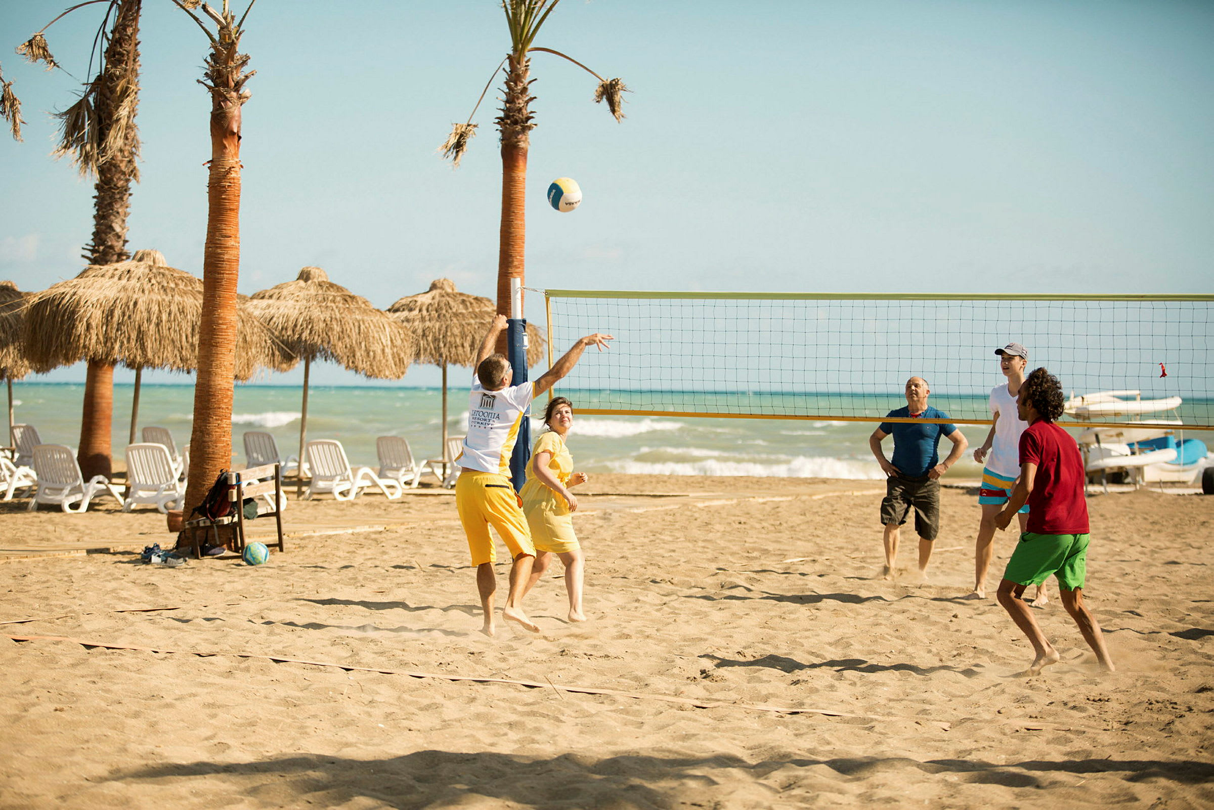 Personer spiller volleyball på en sandstrand med palmer og hav i bakgrunnen. (AI-generert tekst)