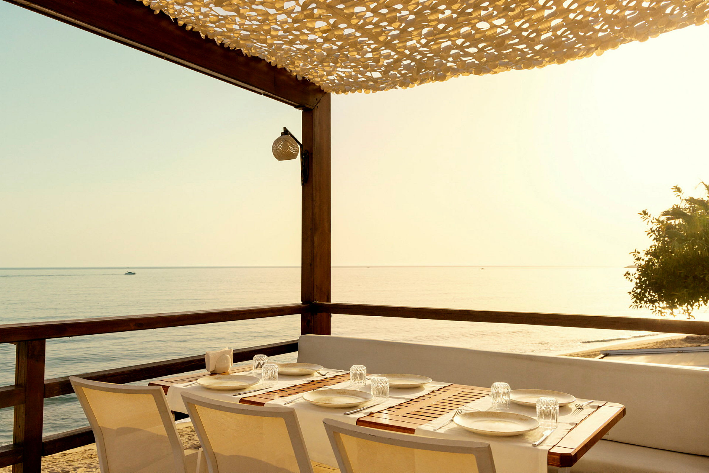 Et bord med borddekking på en terrasse ved havet under en skyggegivende tak. (AI-generert tekst)