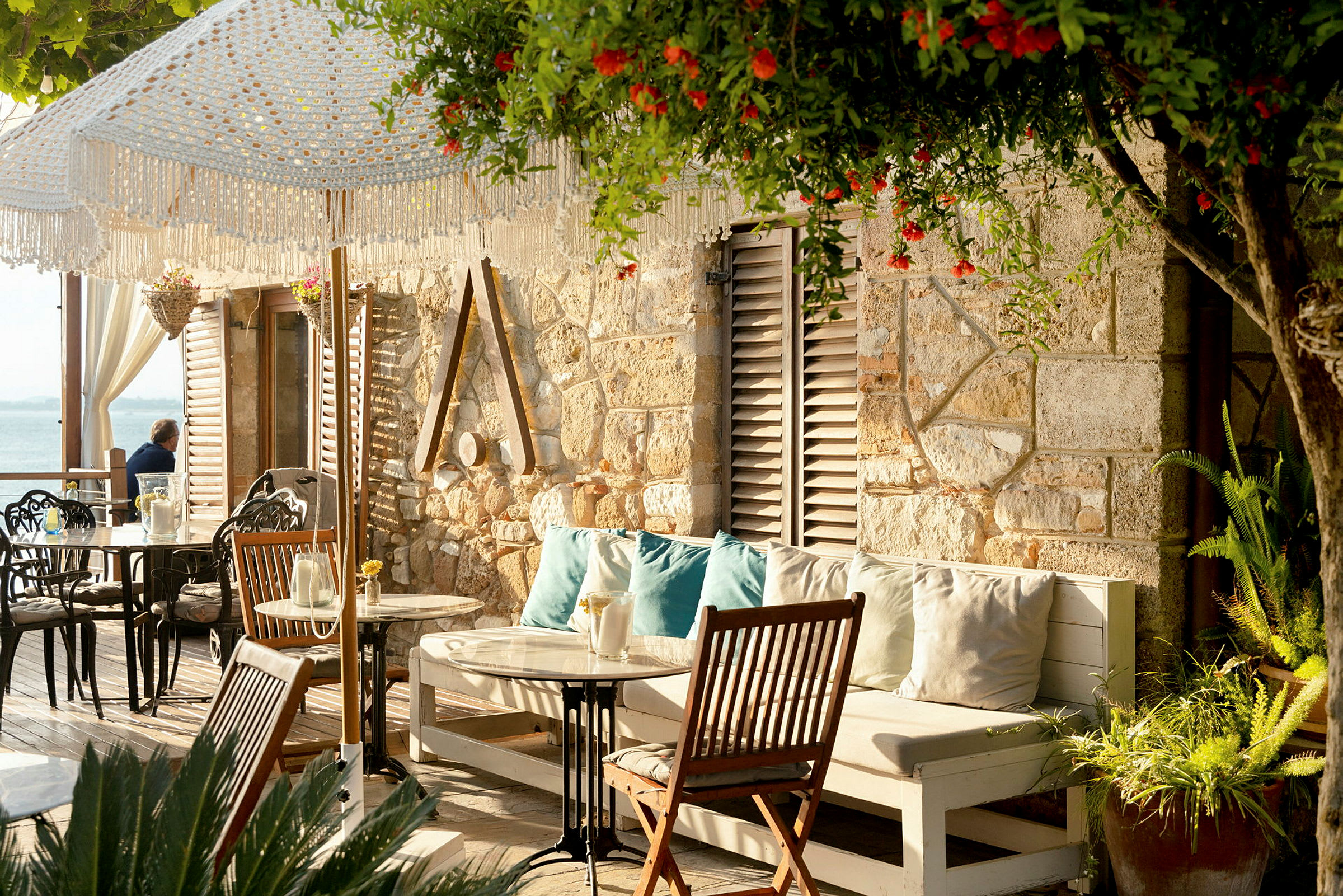 Bilde av en terrasse med sitteplasser, bord og planter ved en steinvegg. Solskinn med lysdekorasjoner og hav i bakgrunnen. (AI-generert tekst)