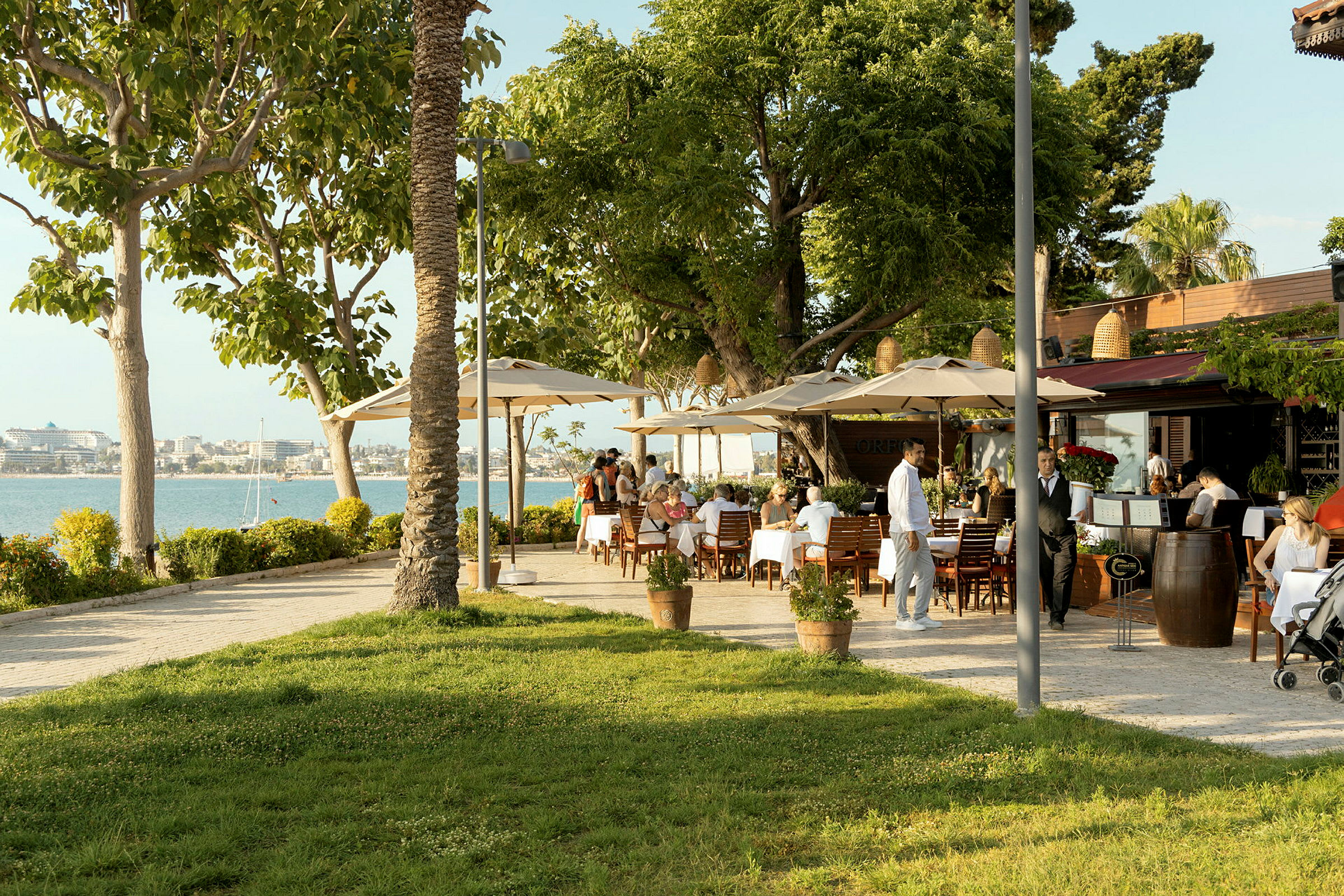 Restaurant med uteservering, bord med hvite duker og grønne trær nær vannet. (AI-generert tekst)