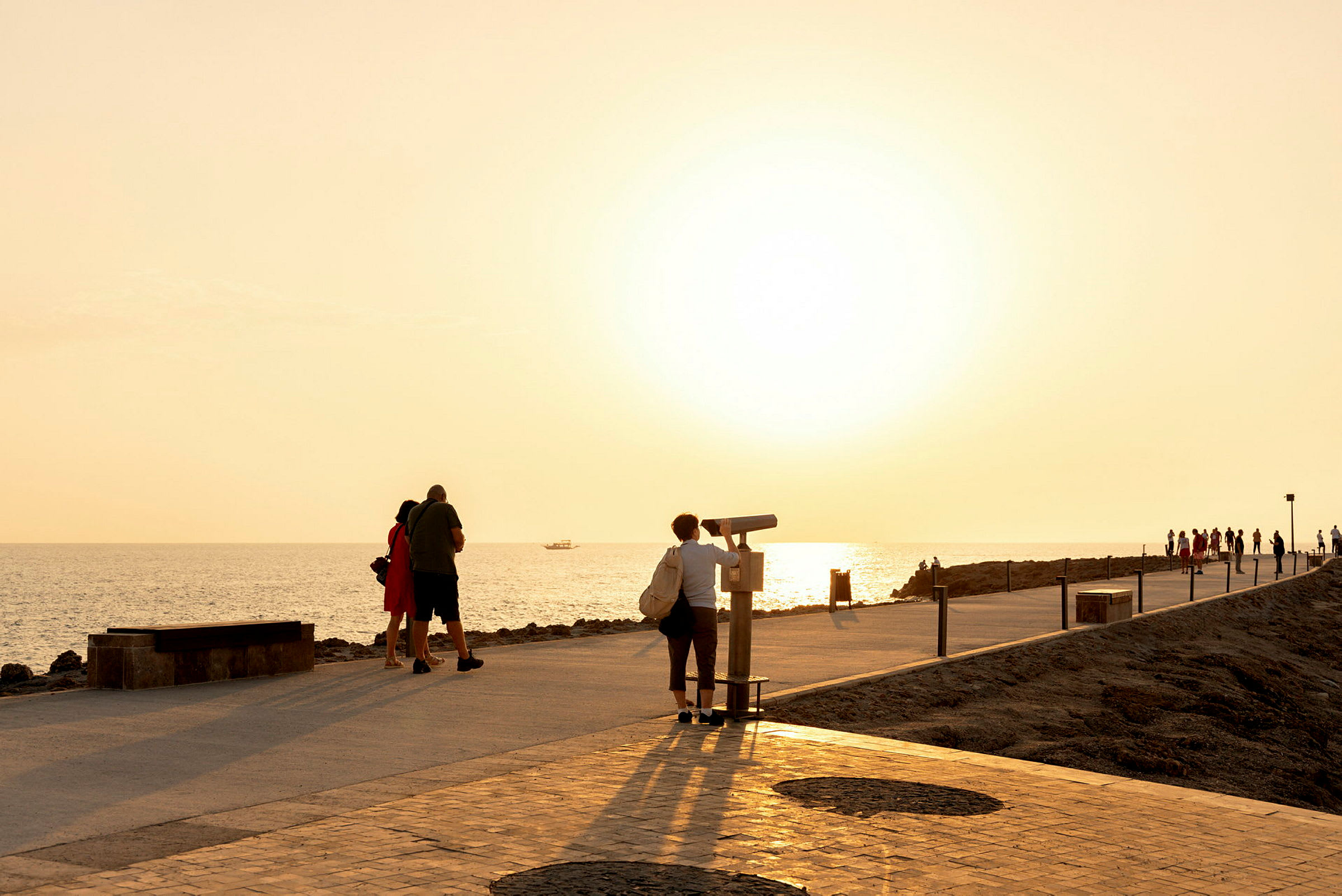 Personer går langs en strandpromenade nær havet i solnedgangen. (AI-generert tekst)