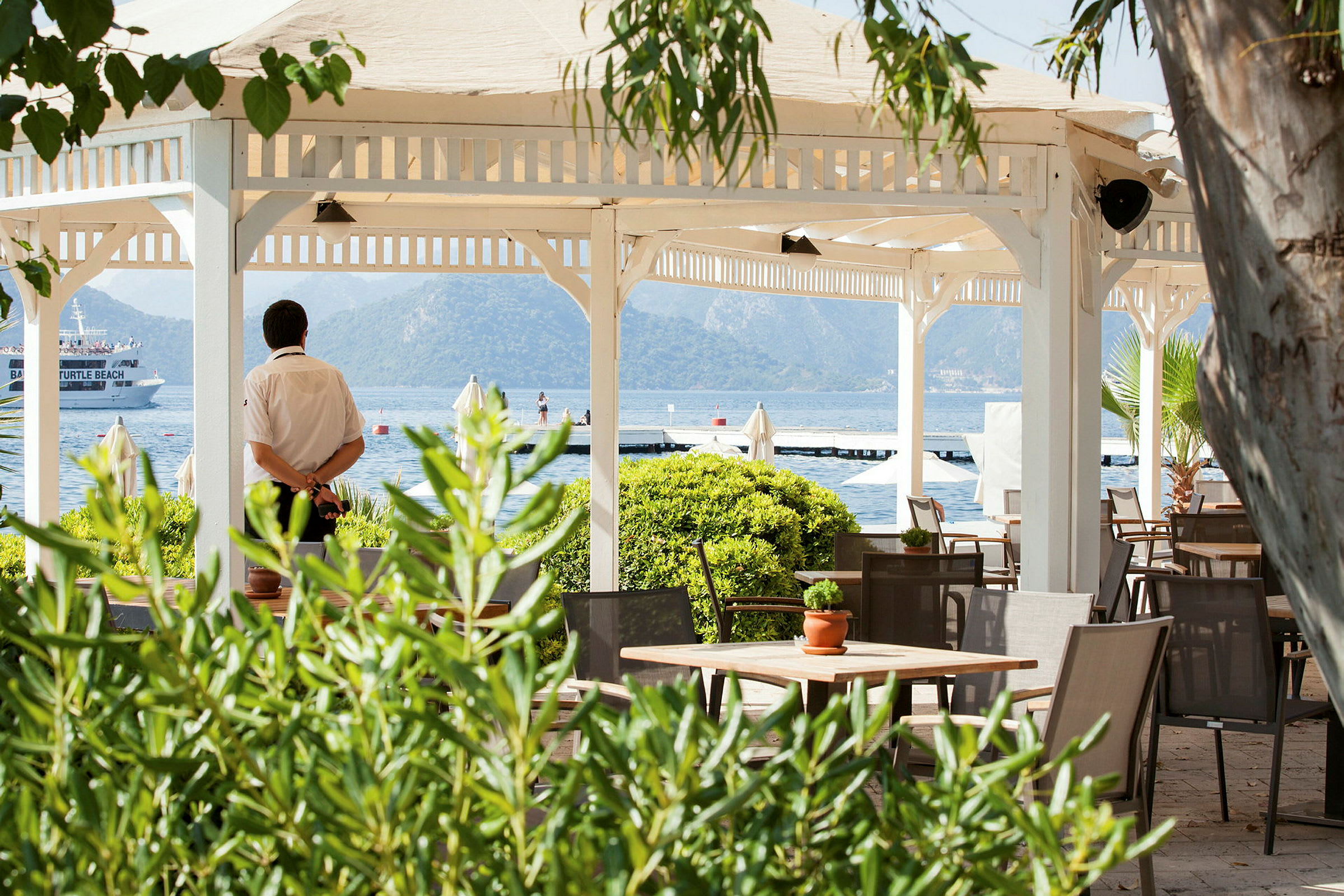 En åpen paviljong med bord og stoler ved havet, omgitt av grøntområder. (AI-generert tekst)