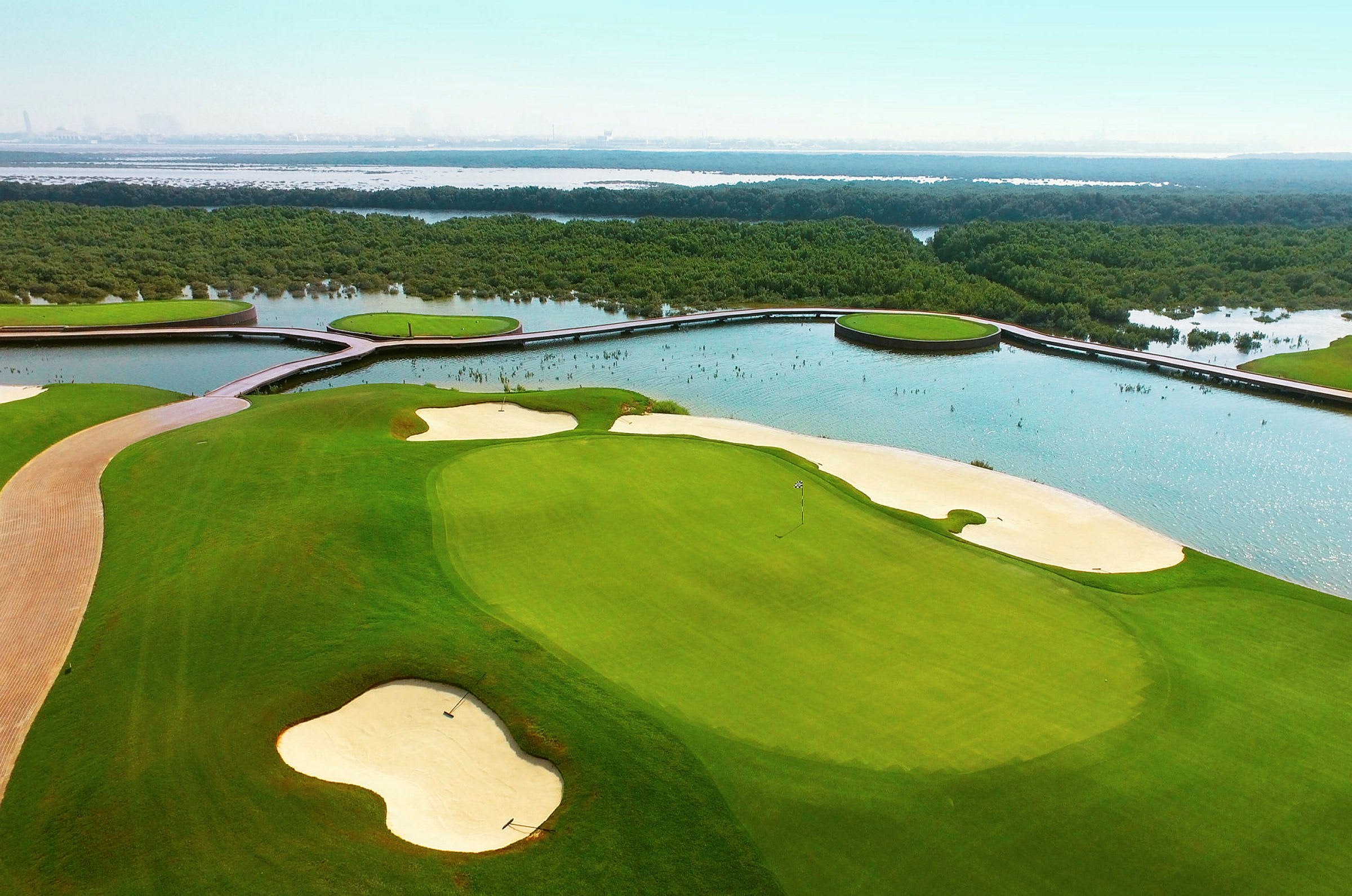 Bildet viser en golfbane med sandbunkere og grønne fairways, omgitt av vann og vegetasjon. (AI-generert tekst)