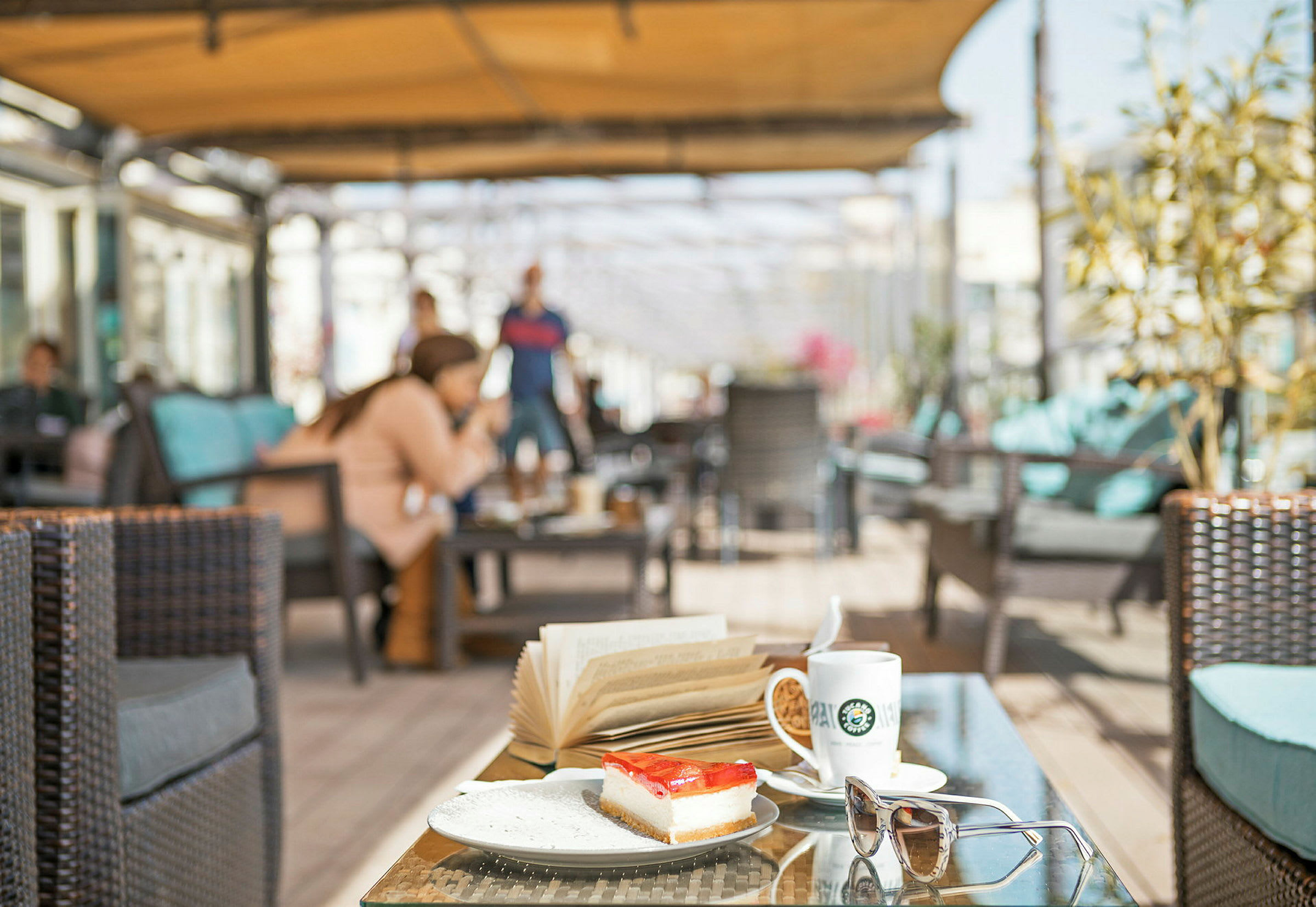 Mat og drikke på et bord med en kaffekopp, en bok og en dessert i en utendørs lounge. (AI-generert tekst)