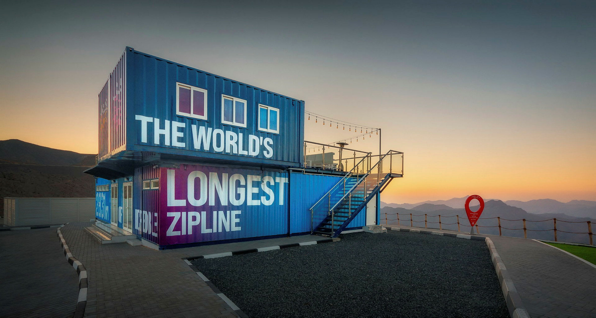 Blå og lilla bygning med teksten 'The World's Longest Zipline' ved solnedgang, omgitt av fjell. (AI-generert tekst)