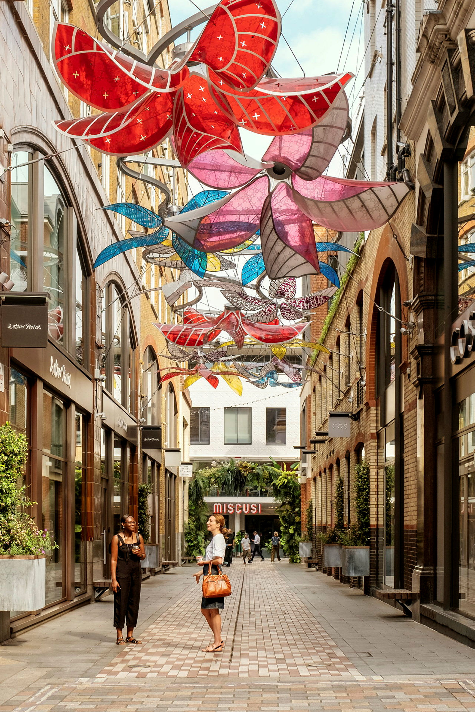 En fargerik dekorasjon i form av blomster henger over en gate med butikker. (AI-generert tekst)