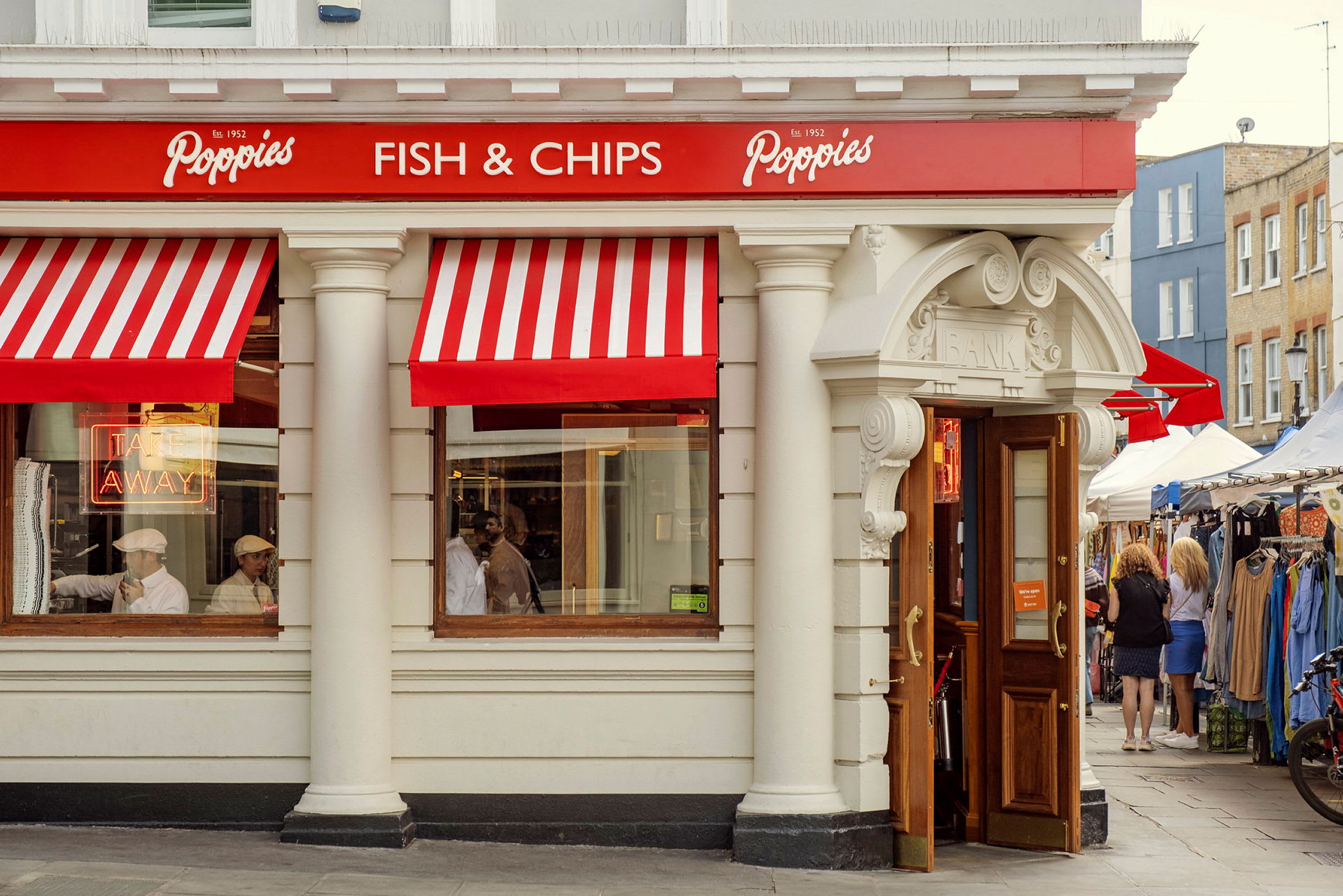 Fasaden til en restaurant med røde og hvite markiser, skiltet 'Fish & Chips', og åpne dører. (AI-generert tekst)