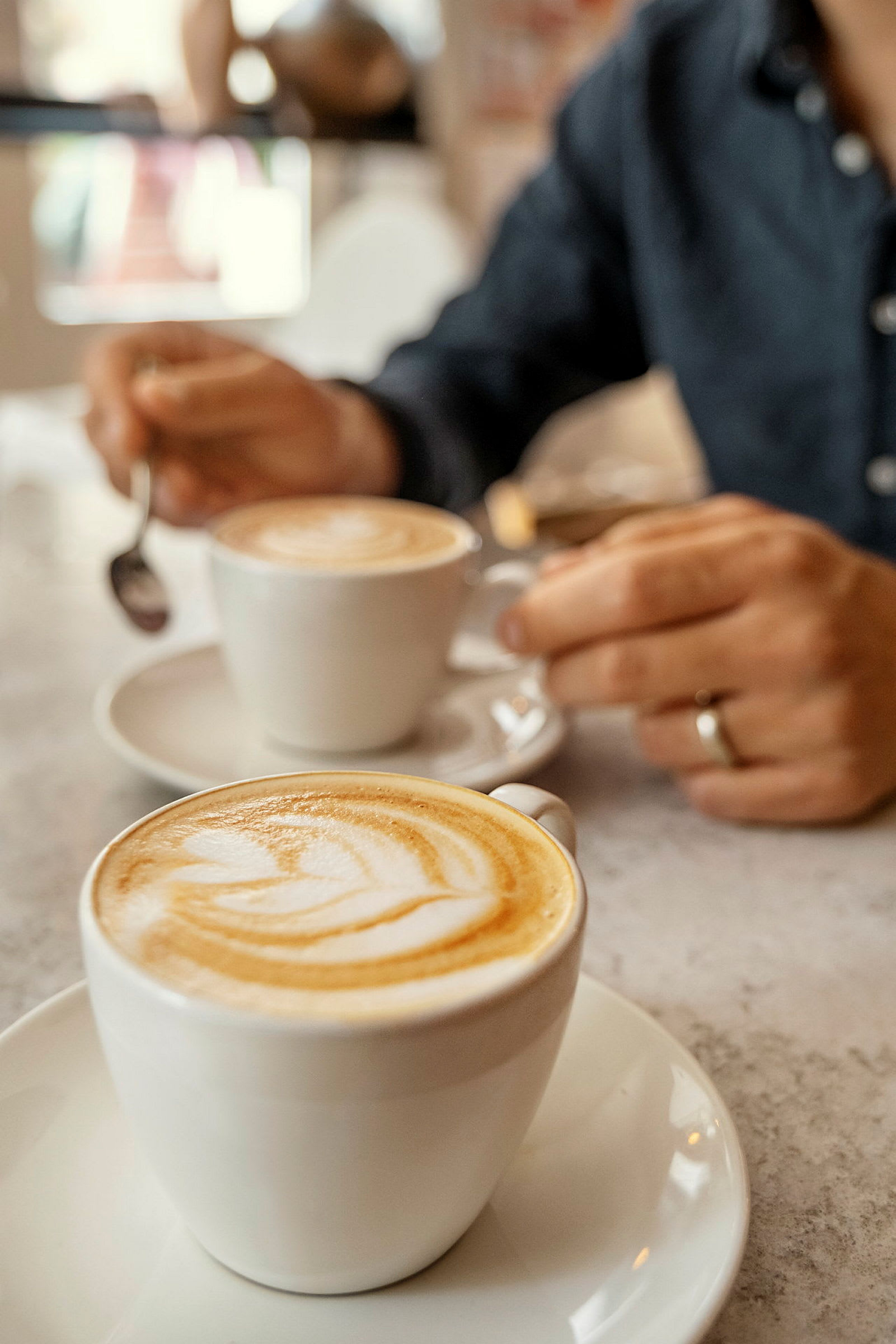To cappuccino i hvite kopper på et bord, med en hånd som holder en skje. (AI-generert tekst)