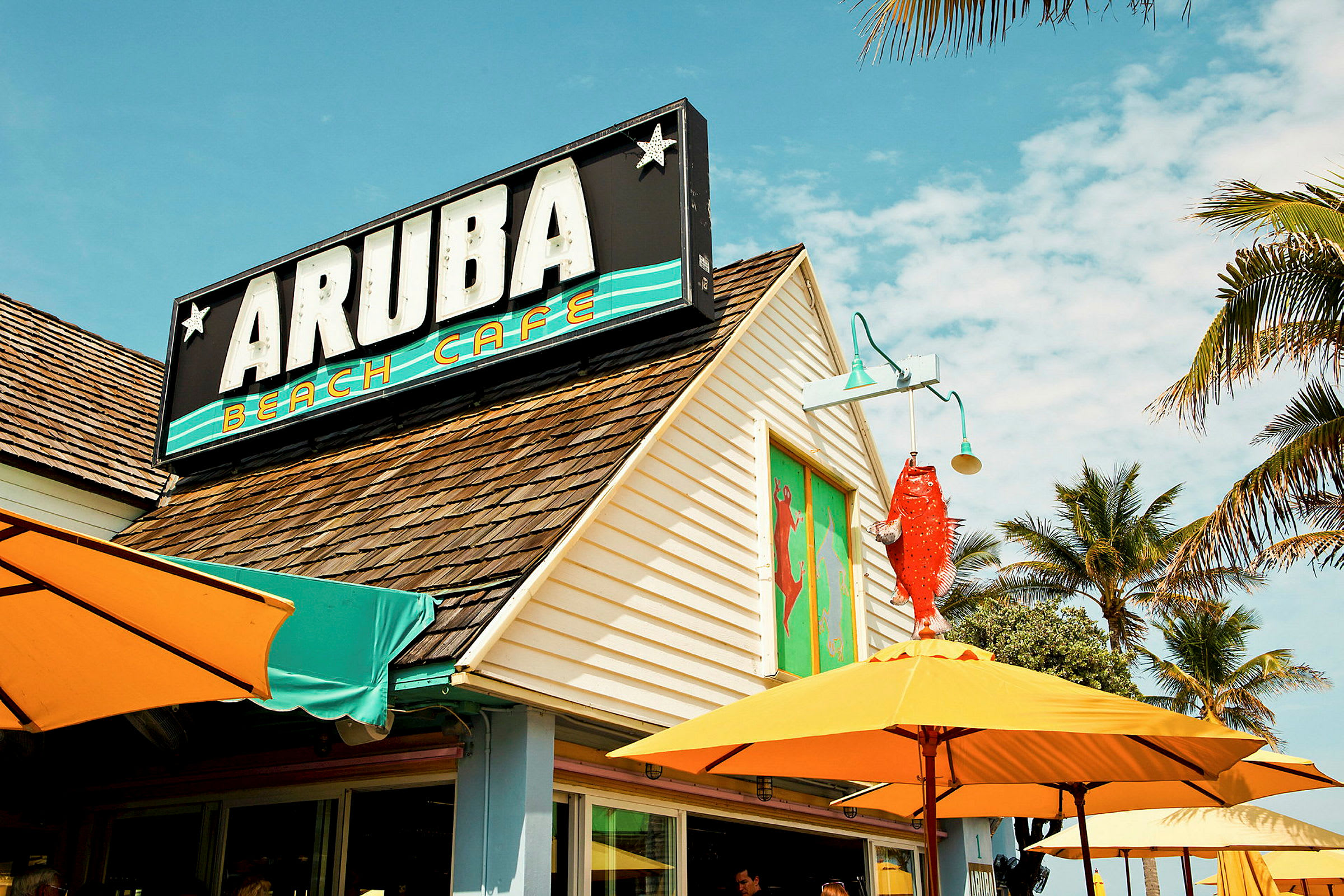 En bygning med et skilt med teksten "Aruba Beach Cafe" og tak med brune takstein. Fargede parasoller og palmer er synlige rundt kafeen. (AI-generert tekst)