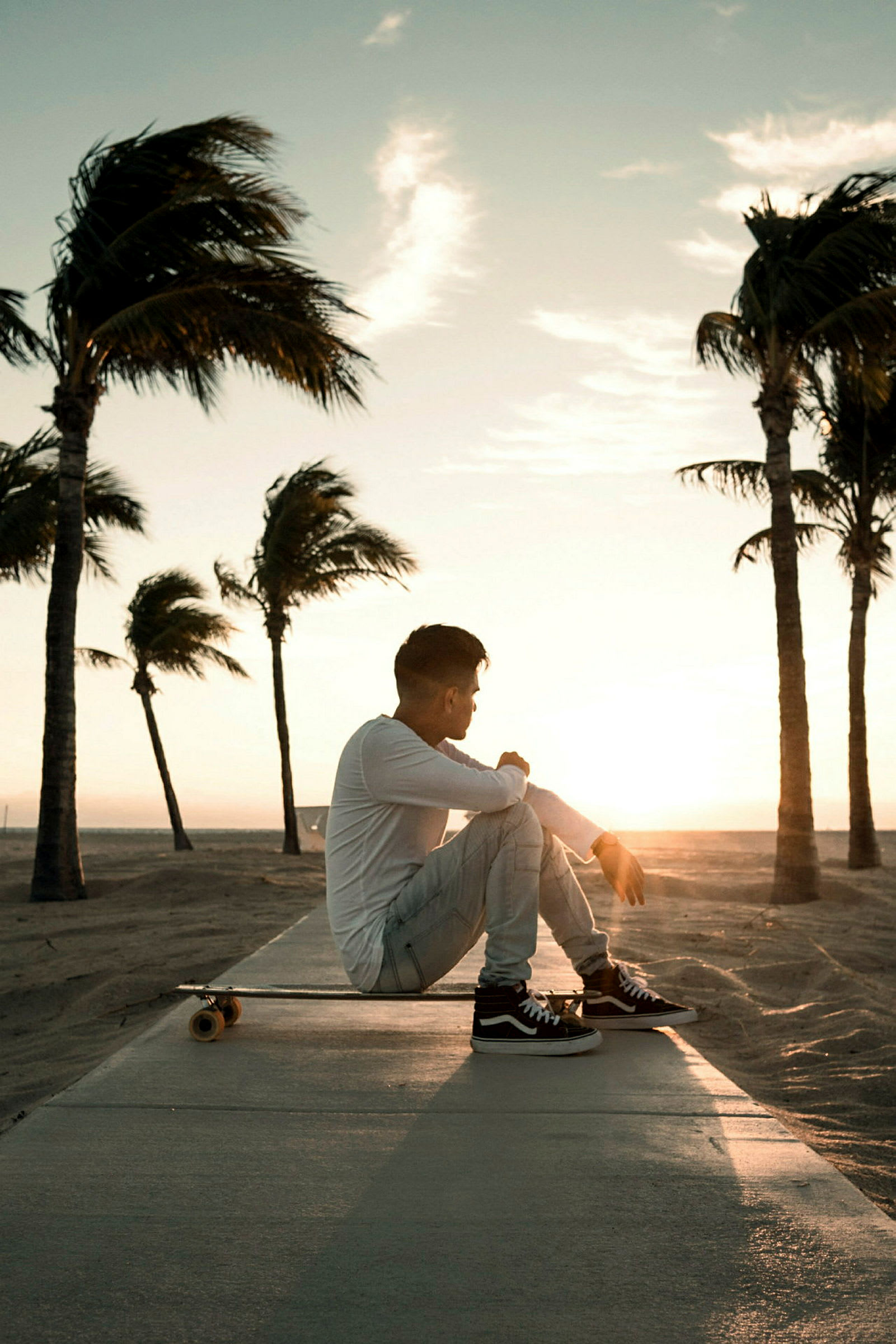 En person sitter på et skateboard ved en strand med palmer i solnedgangen. (AI-generert tekst)
