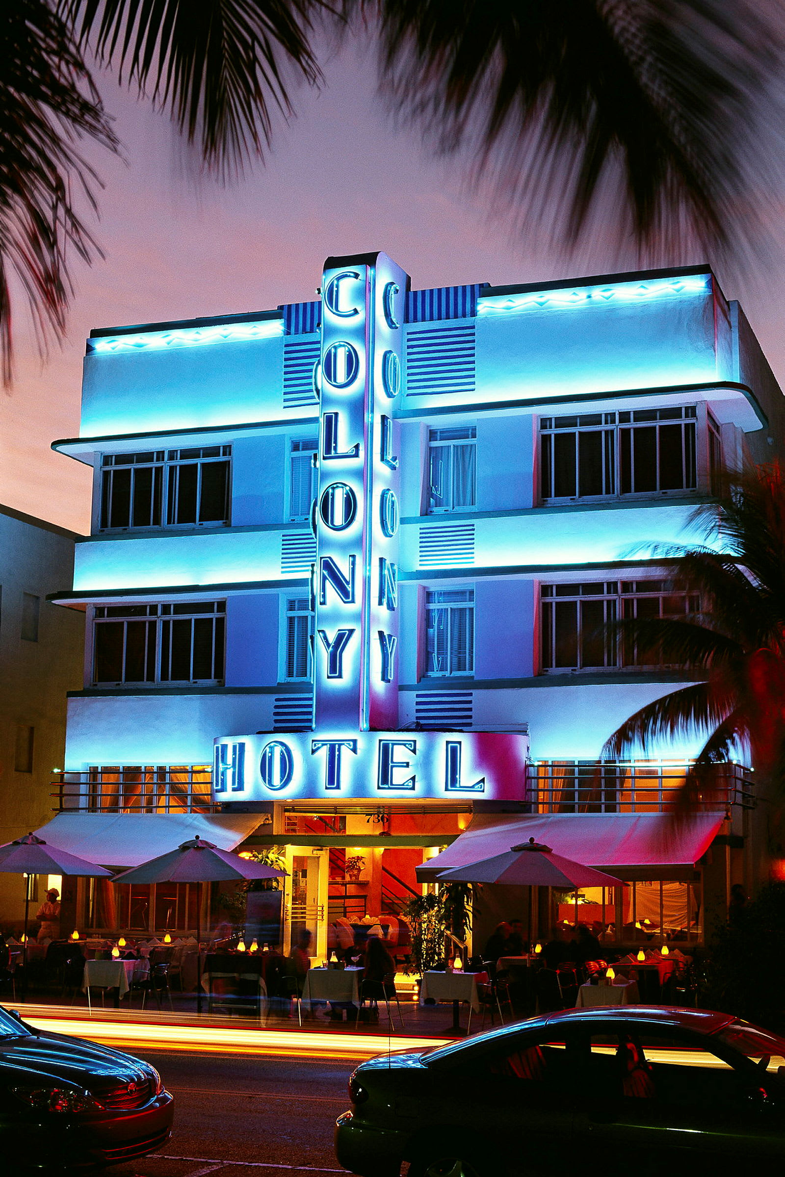Eksteriør av Colony Hotel opplyst i neon om natten med utendørs servering og kjøretøy foran. (AI-generert tekst)