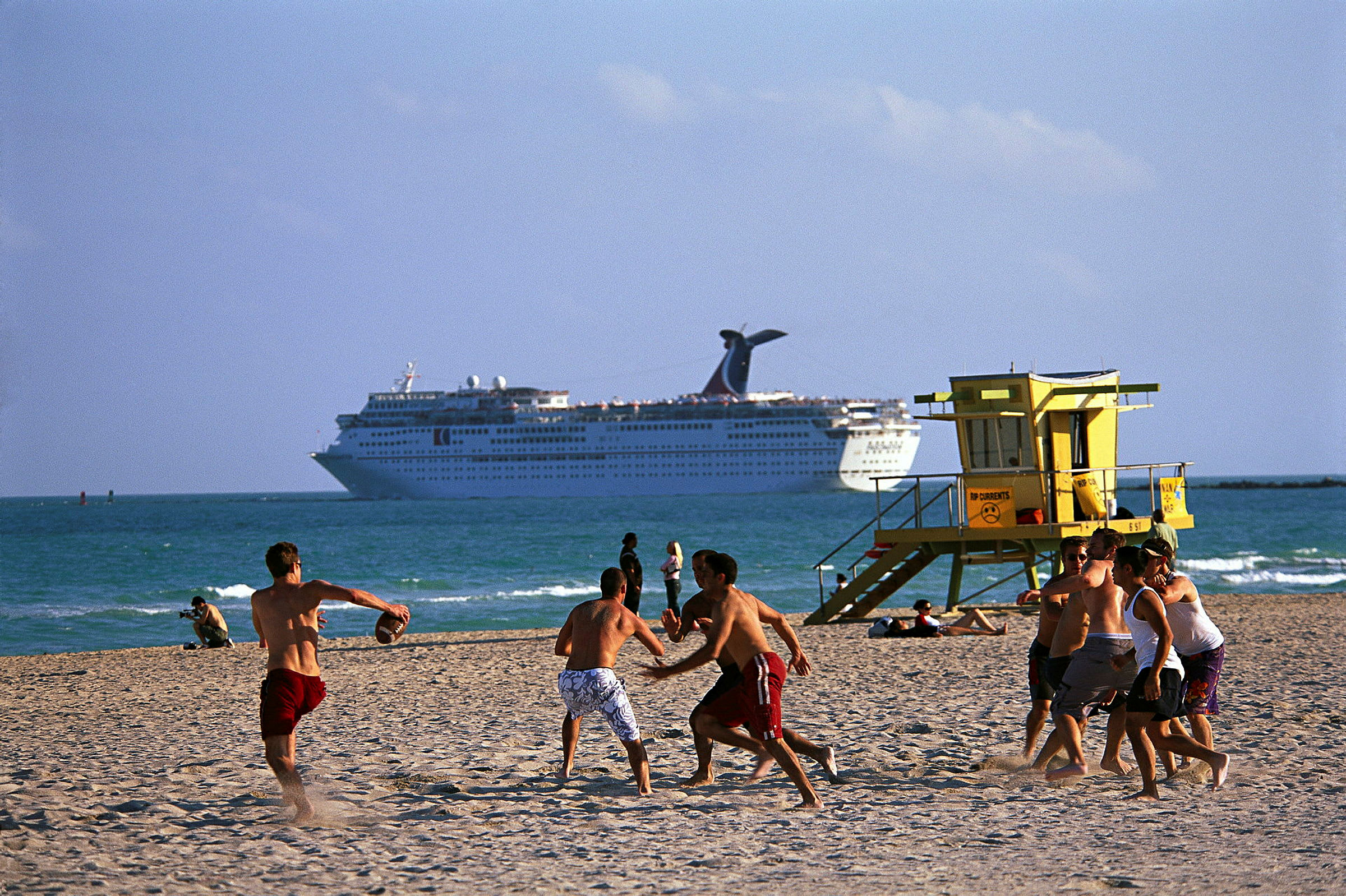 Folk spiller ball på en sandstrand med et cruiseskip i bakgrunnen. (AI-generert tekst)