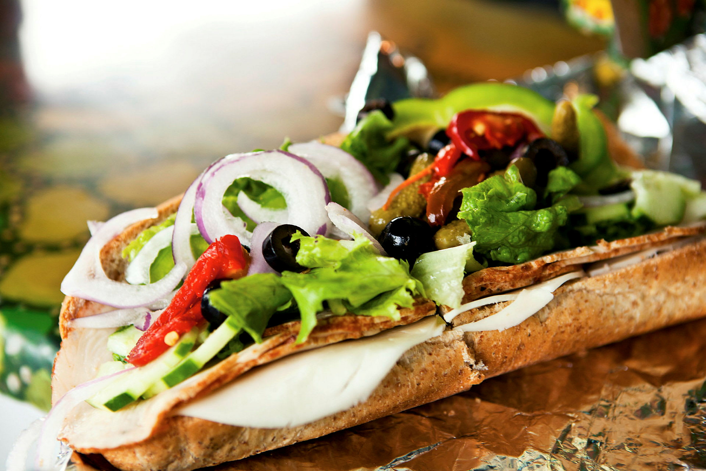 Et nærbilde av en sandwich fylt med salat, ost, rødløk, oliven og paprika på folie. (AI-generert tekst)
