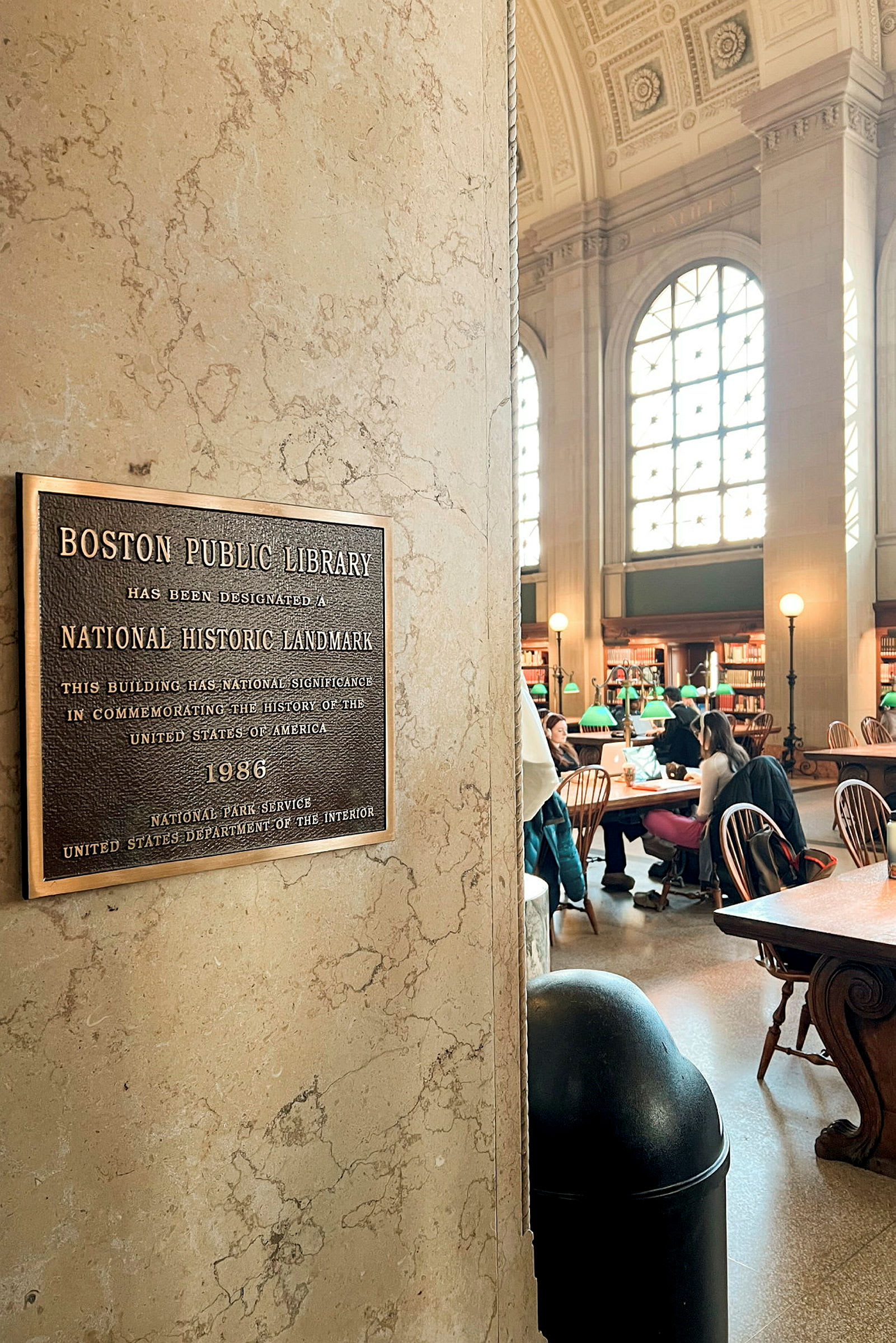 Skilt på en marmorsøyle som forklarer bibliotekets historiske betydning med utforskende besøkende og belysning i bakgrunnen. (AI-generert tekst)