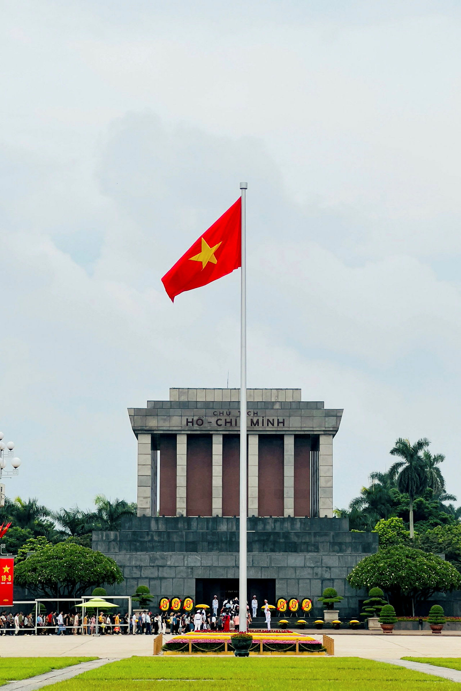 Et stort mausoleum med teksten 'Ho Chi Minh' på fasaden, foran hvilket det vietnamesiske flagget er reist på en flaggstang. Folk samler seg foran bygningen som er omgitt av trær og plener. (AI-generert tekst)