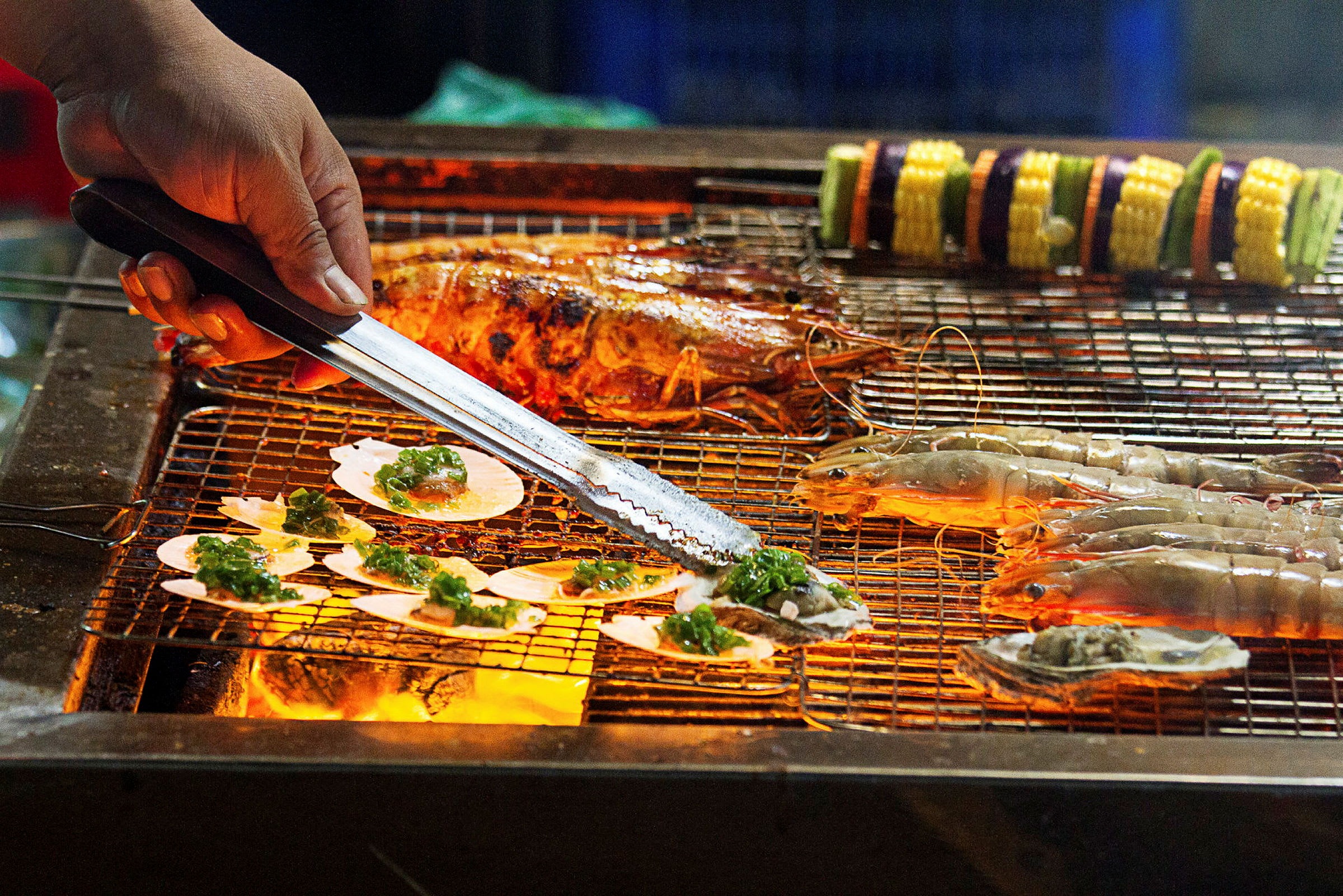 En grill med skalldyr og grønnsaksspyd, der en hånd håndterer maten med tang. (AI-generert tekst)