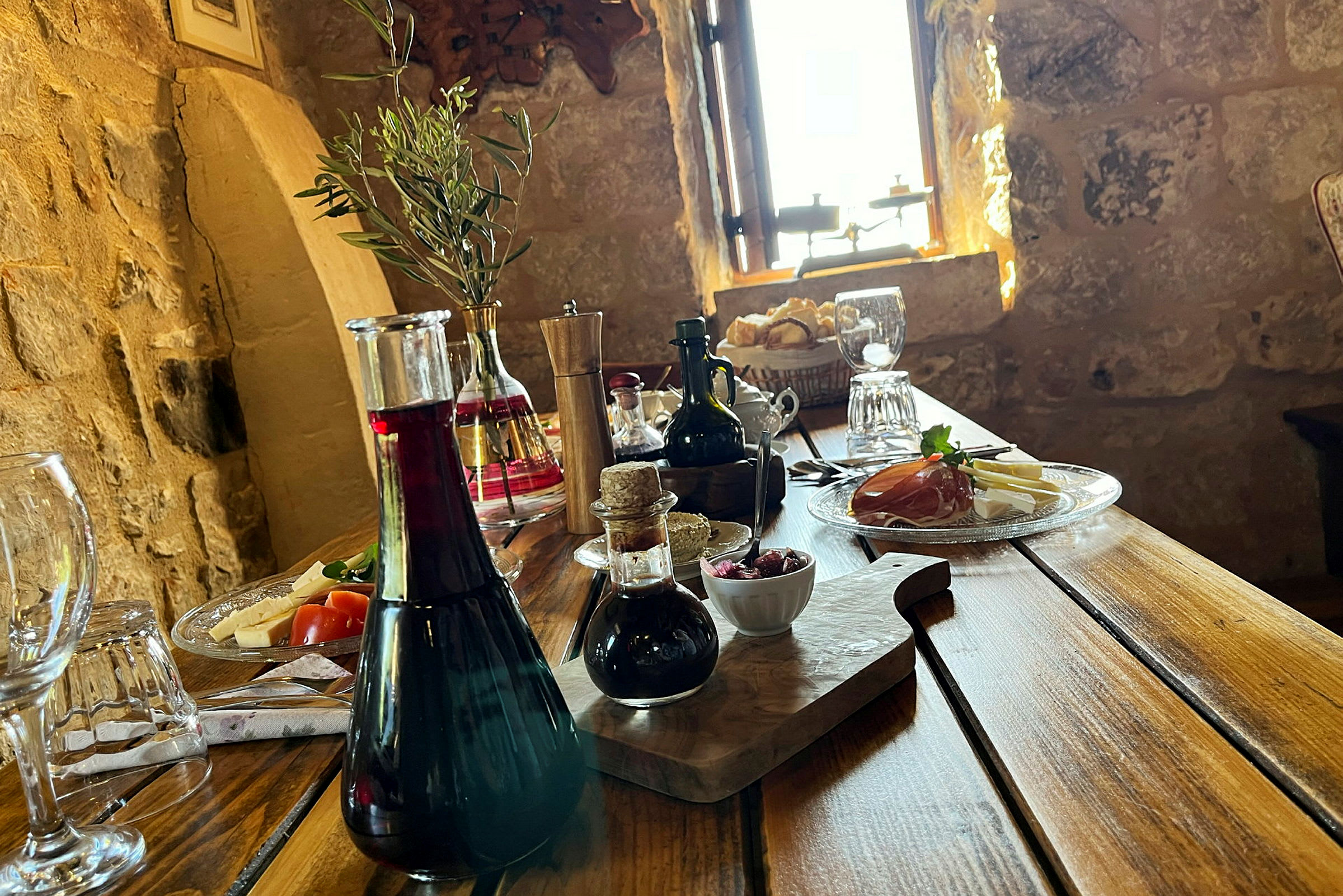 Et rustikt trebord med forskjellige serveringer, inkludert olivenolje, vin, brød og spekemat, samt dekorasjoner i et eldre miljø. (AI-generert tekst)