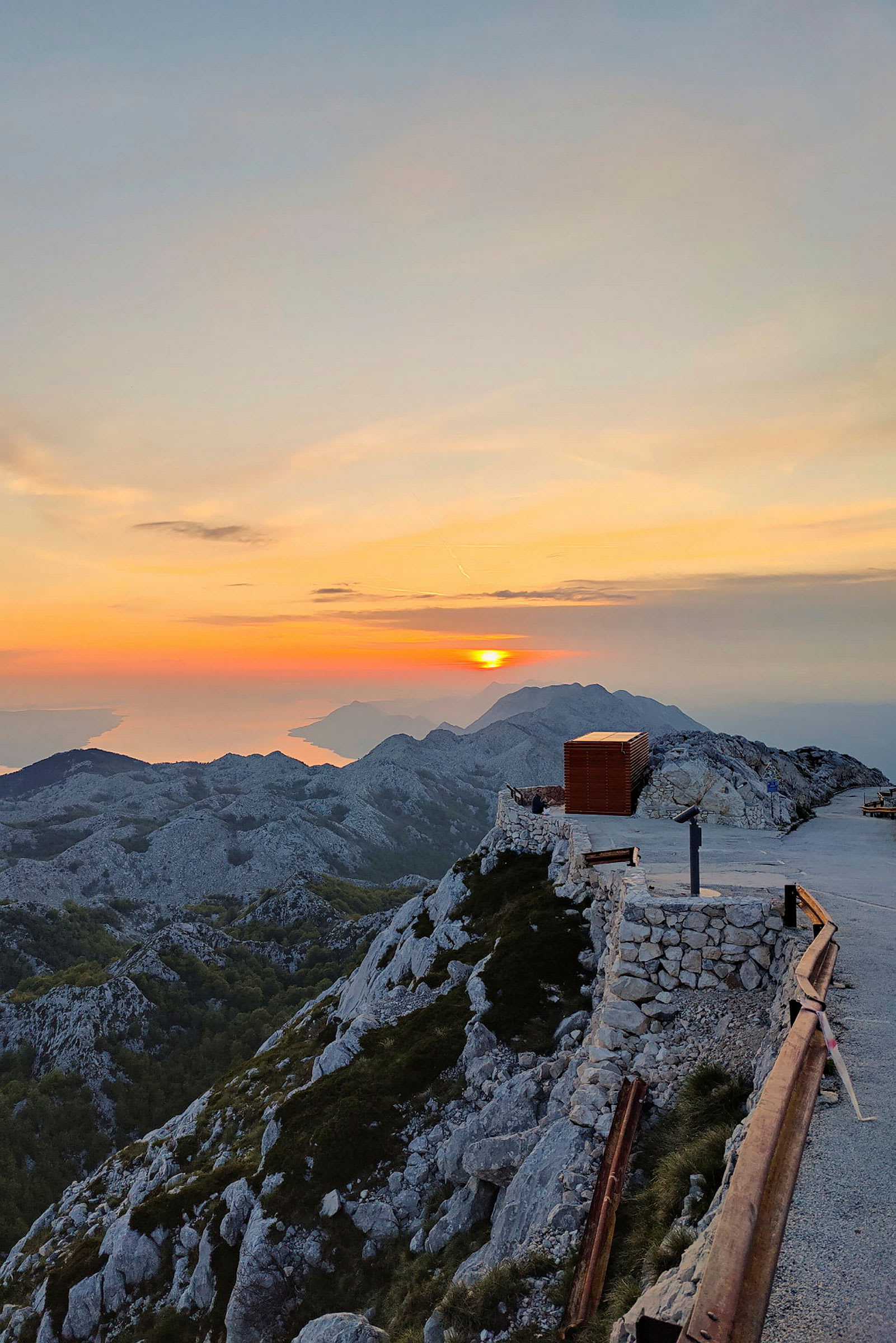 Bilde av en fjelltopp under solnedgang med steinmur, en gangvei og et rekkverk i forgrunnen. (AI-generert tekst)
